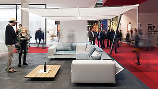 Canapé-lit gris à l’intérieur du stand de la foire, avec des coussins de dossier réglables et un paysage marin de coucher de soleil encadré par des tiges blanches verticales.
