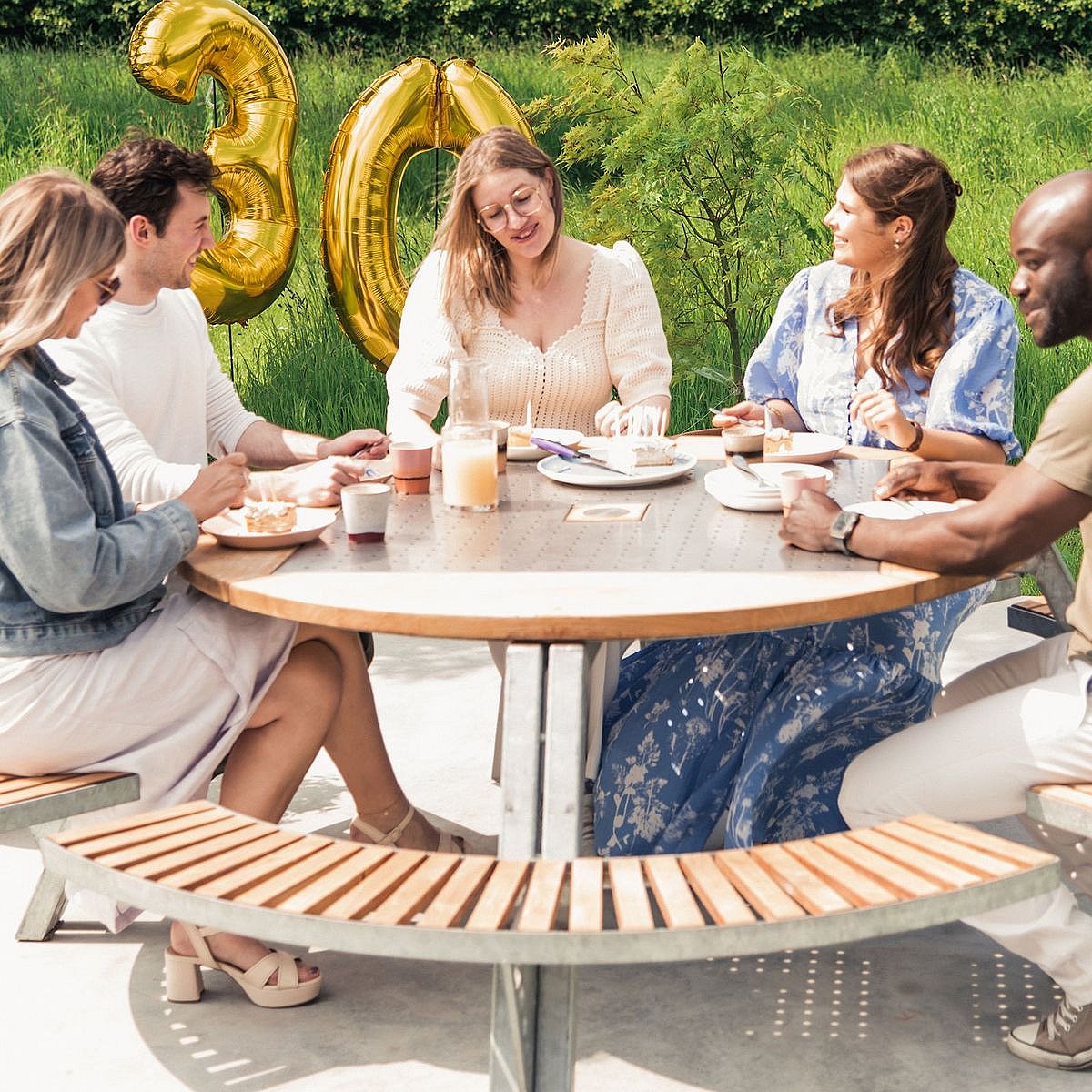 Vijf mensen zitten buiten rond een ronde Gargantua-picknicktafel en delen taart en drankjes, met grote gouden ballonnen met het cijfer '30' erachter om het 30-jarig bestaan ​​van de tafel te vieren.