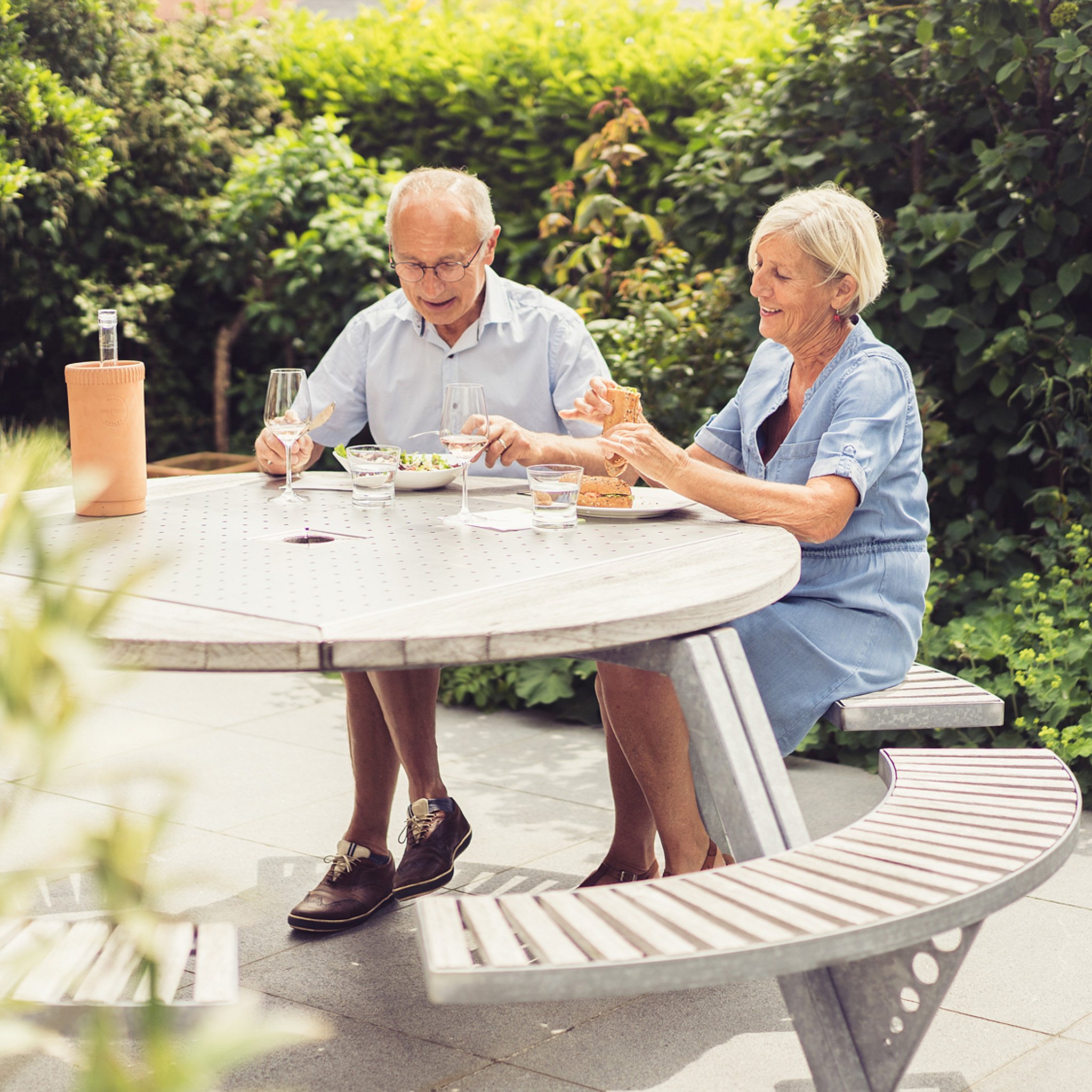 Un couple âgé est assis ensemble à une table ronde de pique-nique Gargantua sur une terrasse ensoleillée, profitant d’un déjeuner avec des verres de vin et de la verdure tout autour.