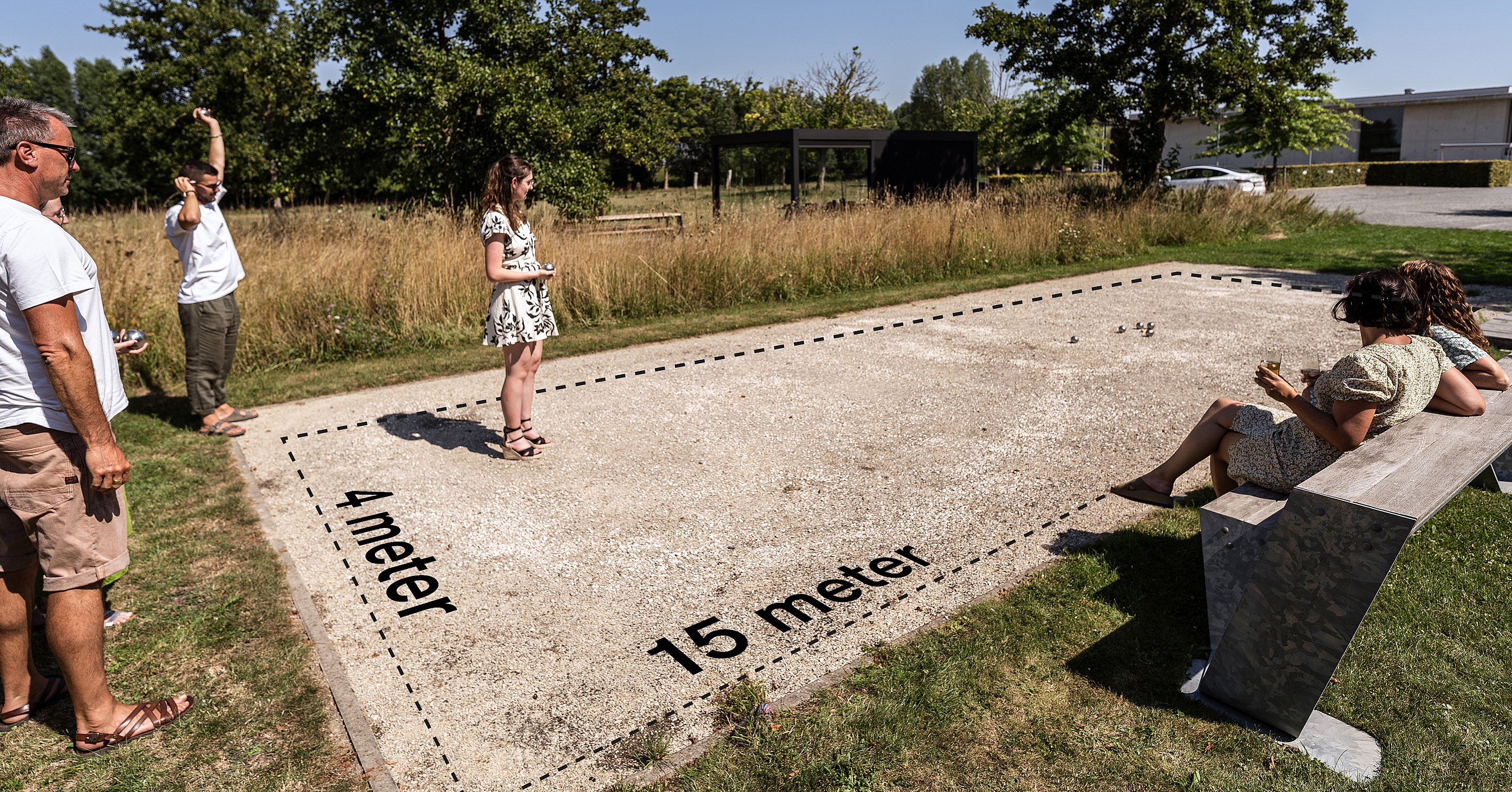 Weitwinkelansicht eines Pétanque-Platzes mit gestrichelten Maßlinien, die eine 4 x 15 Meter große Spielfläche markieren, wobei eine Frau auf dem Platz steht und eine andere Person auf einer Bank sitzt.