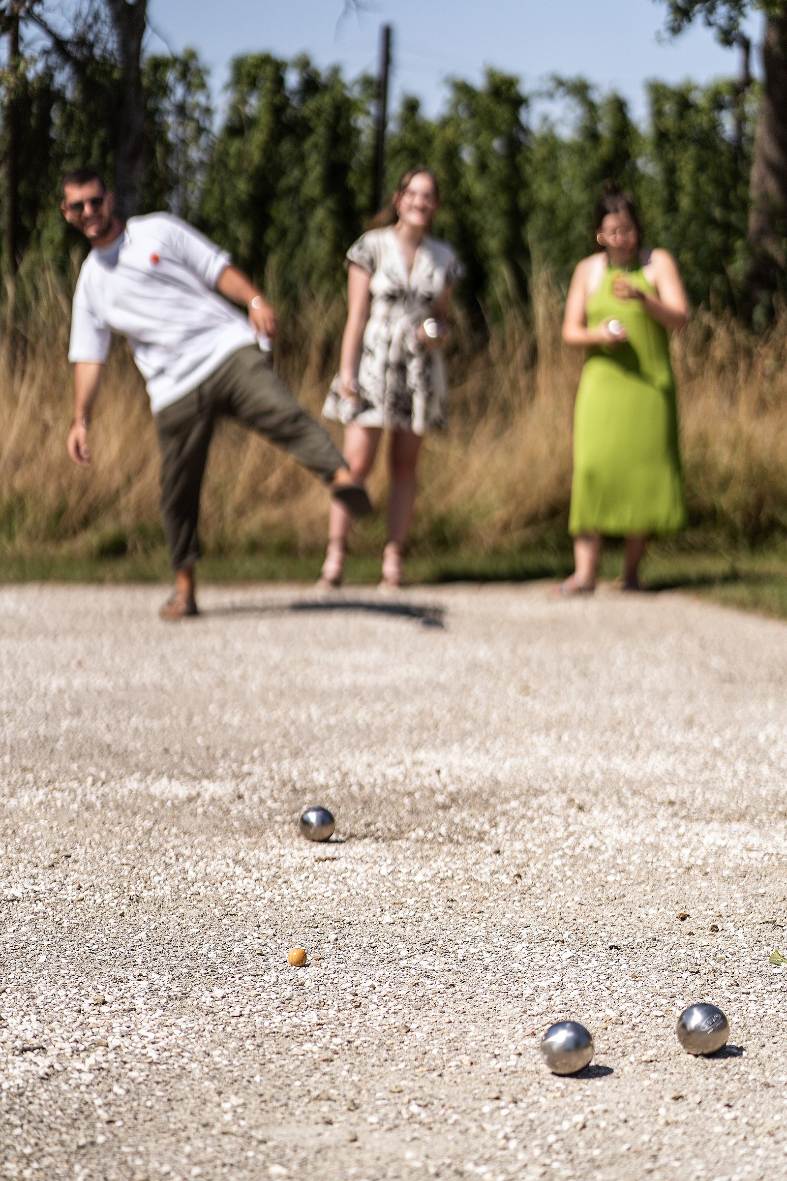 Mann im weißen T-Shirt wirft einen Pétanque Boule mit angehobenem Bein, während zwei Teamkollegen von hinten zusehen.