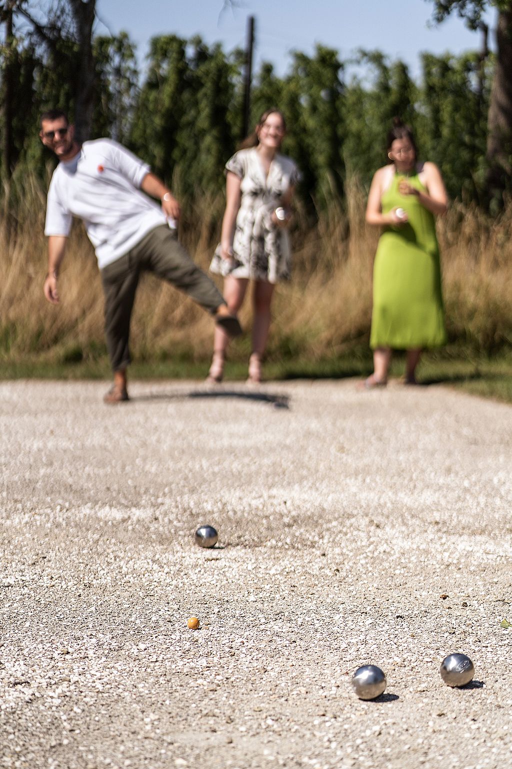 Mann im weißen T-Shirt wirft einen Pétanque Boule mit angehobenem Bein, während zwei Teamkollegen von hinten zusehen.