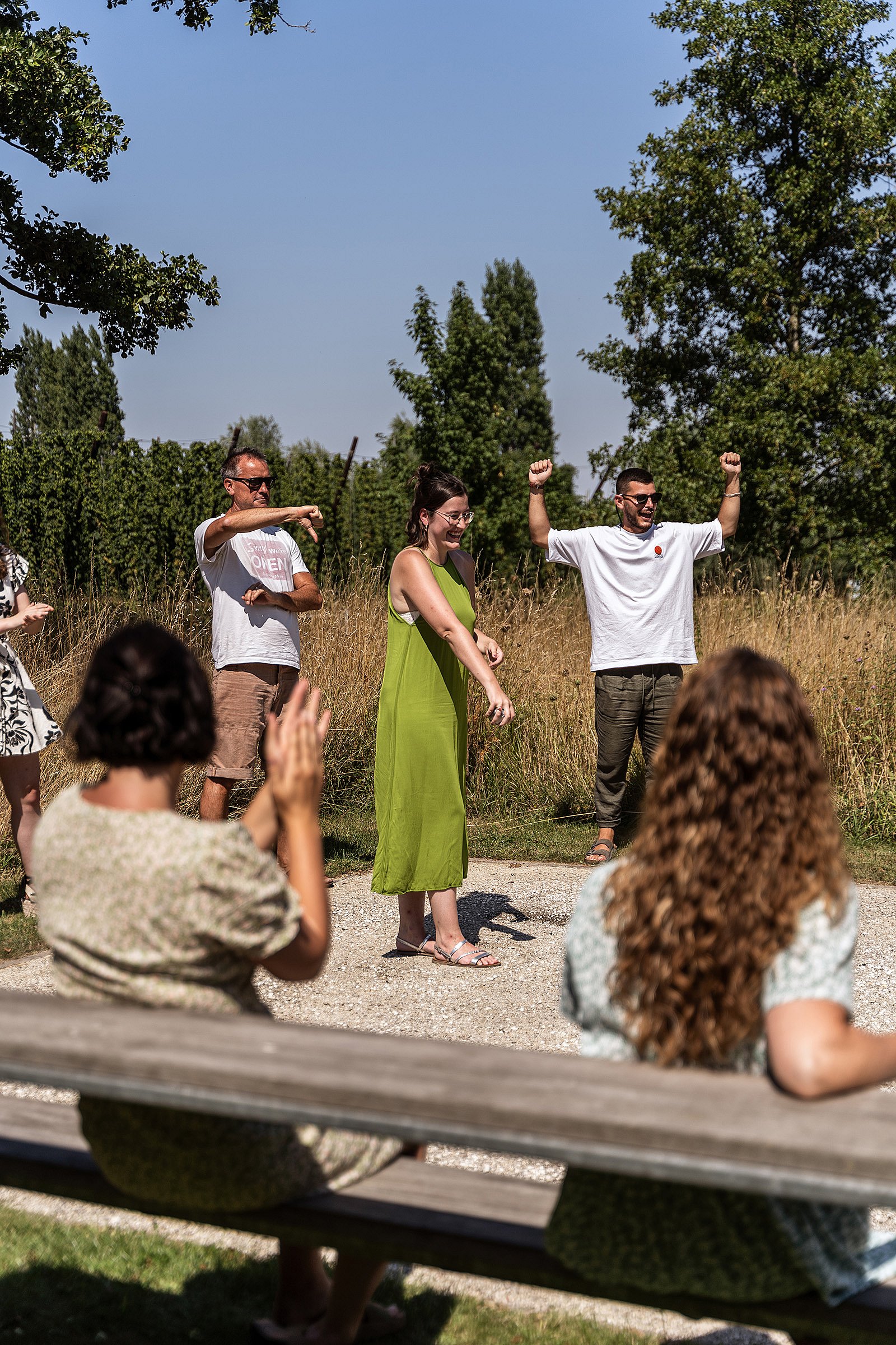 Eine Gruppe von Spielern reagiert auf den jüngsten Wurf auf dem Pétanque-Platz, wobei eine Frau zeigt und andere im Hintergrund jubeln.