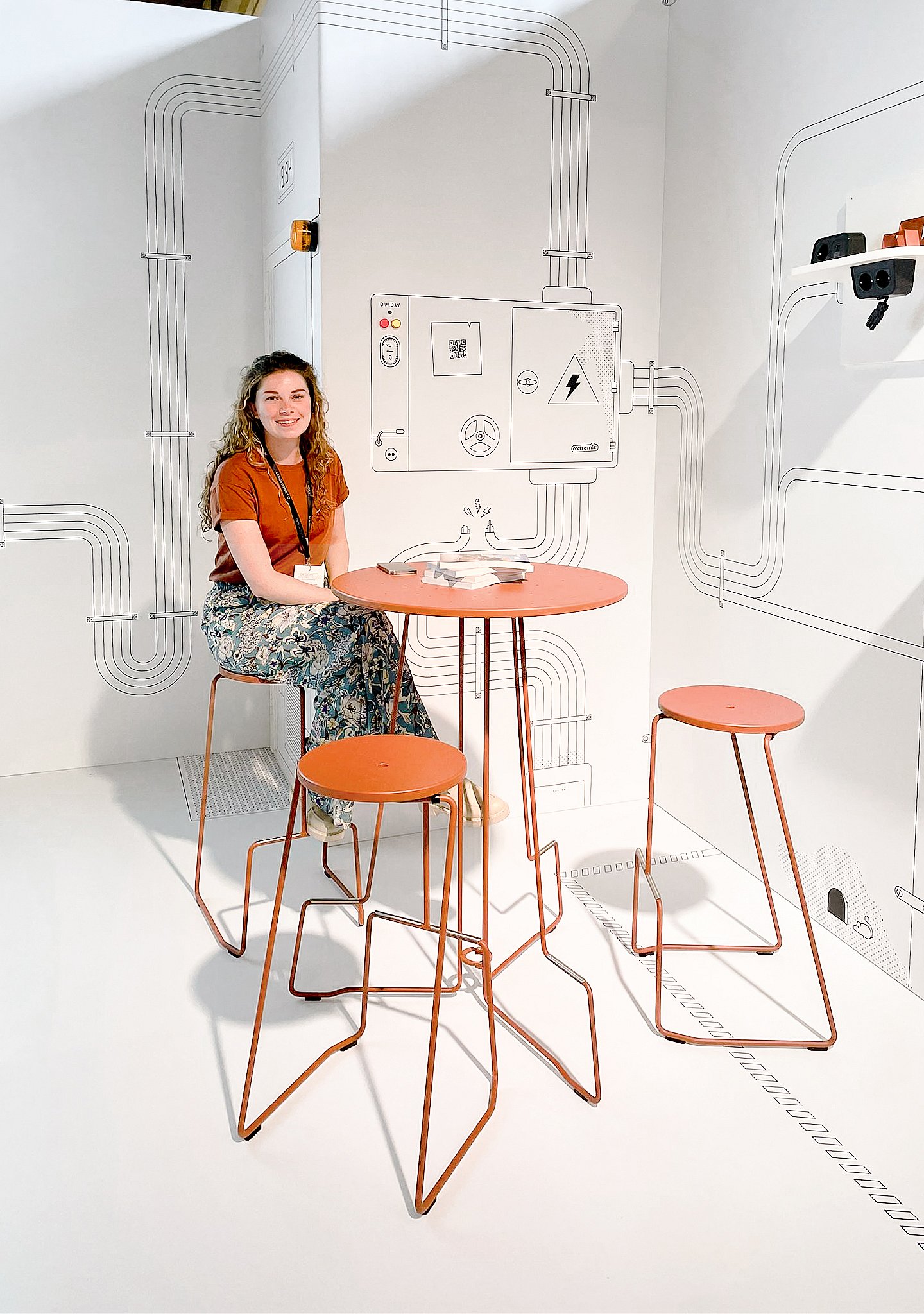 Une femme est assise à une petite table haute orange avec des tabourets assortis, entourée de murs blancs imprimés de dessins de circuits et d'icônes d'énergie.