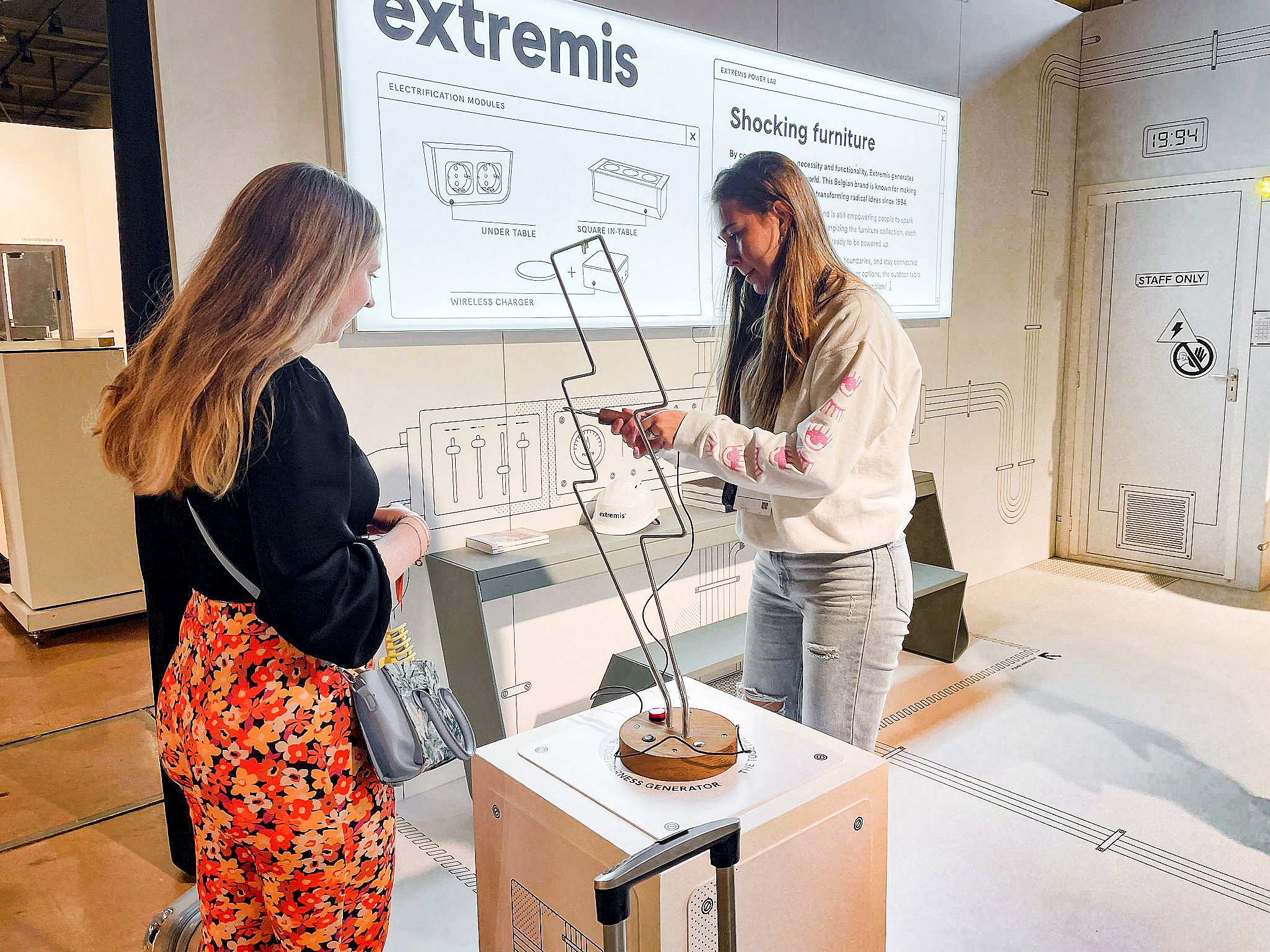 Deux femmes se tiennent près d'un présentoir sur piédestal, observant un objet en forme de zigzag devant un grand panneau mural intitulé « Mobilier électrifiant ».