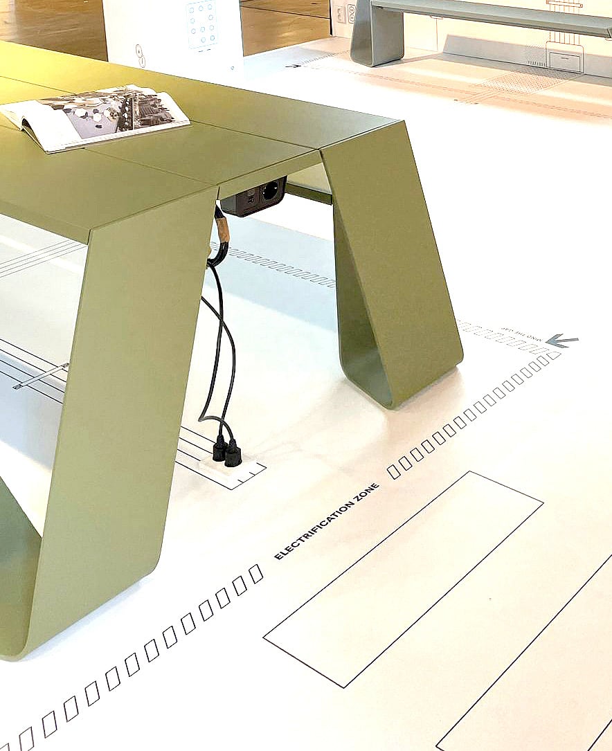 Détail du mobilier vert plié au-dessus des graphismes imprimés au sol, soulignant la pureté des lignes du design et le thème technique de l'exposition.