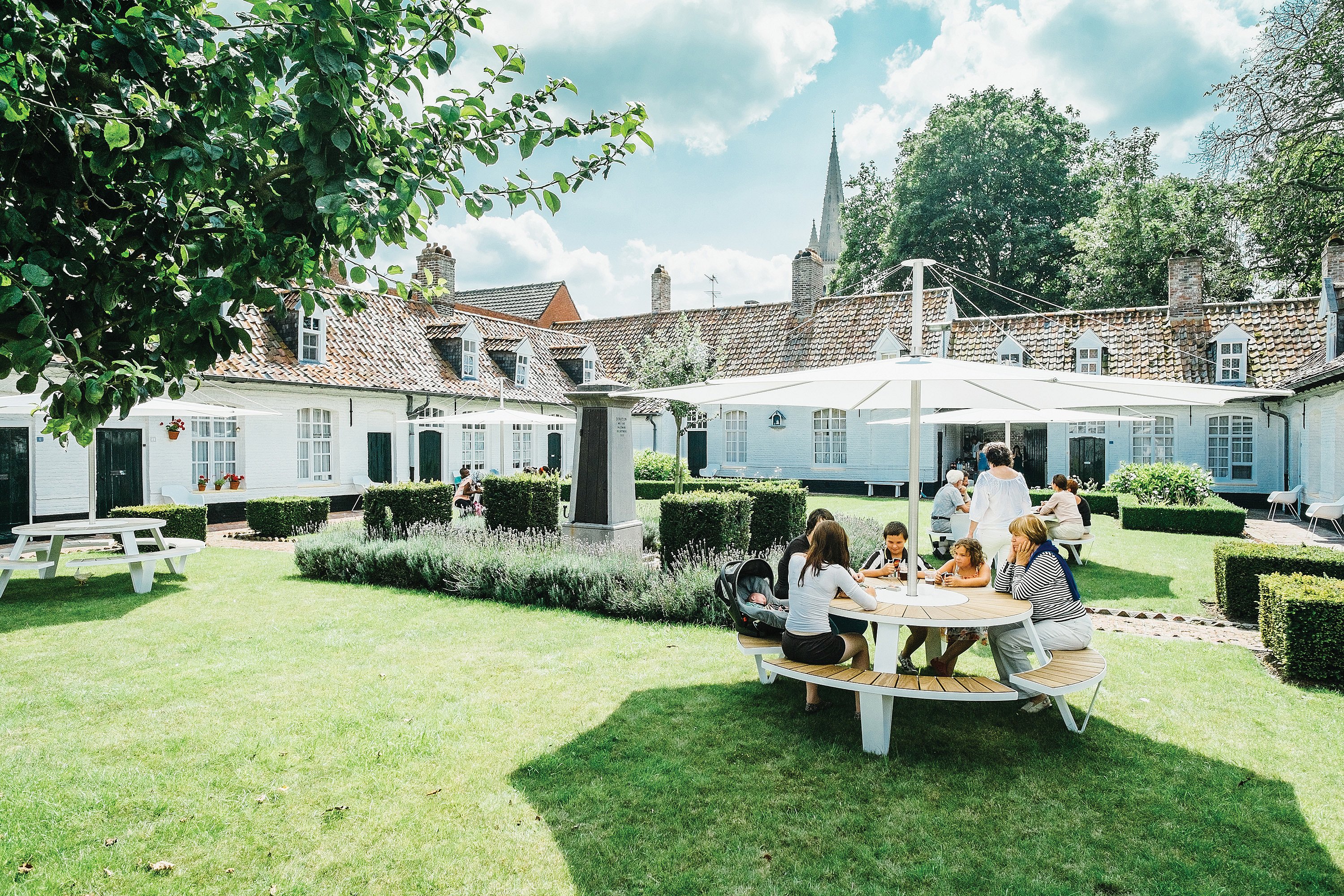 Round Pantagruel picnic table with integrated curved benches and a large white central parasol, used by a family in a sunny courtyard surrounded by white buildings and clipped hedges.