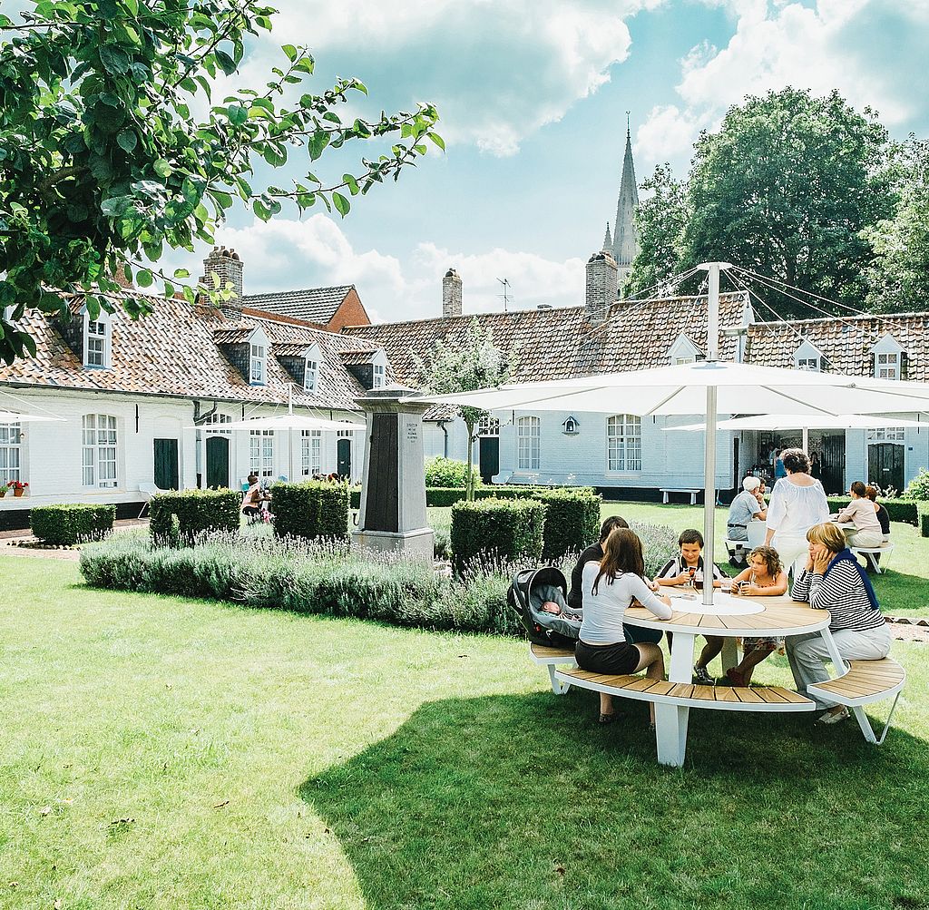 Round Pantagruel picnic table with integrated curved benches and a large white central parasol, used by a family in a sunny courtyard surrounded by white buildings and clipped hedges.