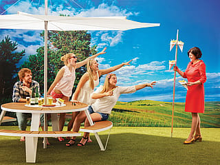 our young adults sitting around a Pantagruel table under a wide white parasol, reaching playfully toward a woman in a coral dress holding a tall stand of cone-shaped snacks.
