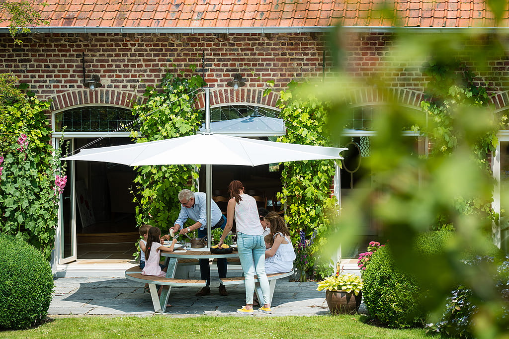 Family gathered around a Pantagruel table on a stone terrace outside a brick house covered with climbing greenery, sharing a meal beneath a white parasol.