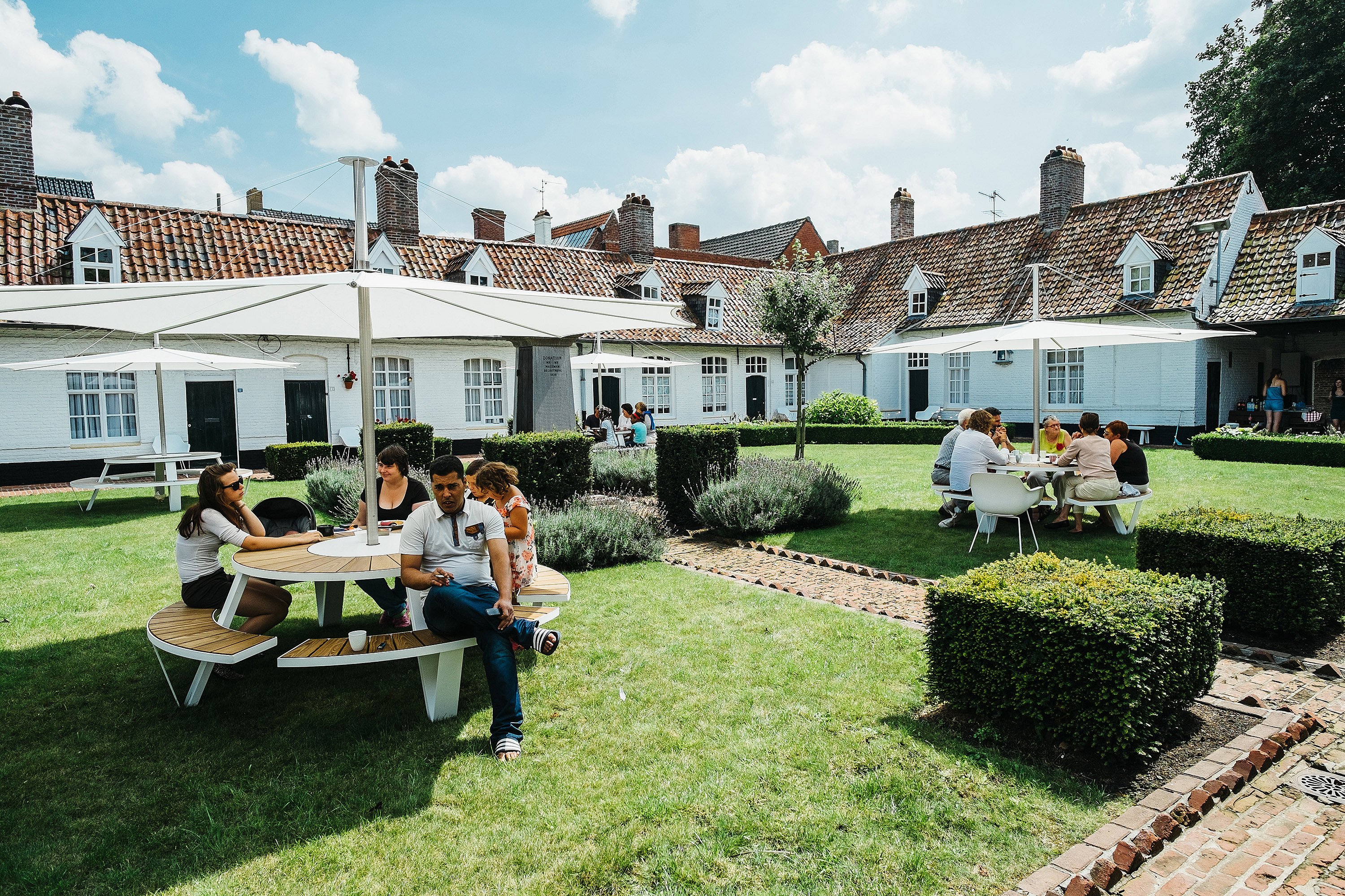 Several groups seated at Pantagruel tables with white parasols in a large grassy courtyard, with white brick buildings and tiled roofs in the background.