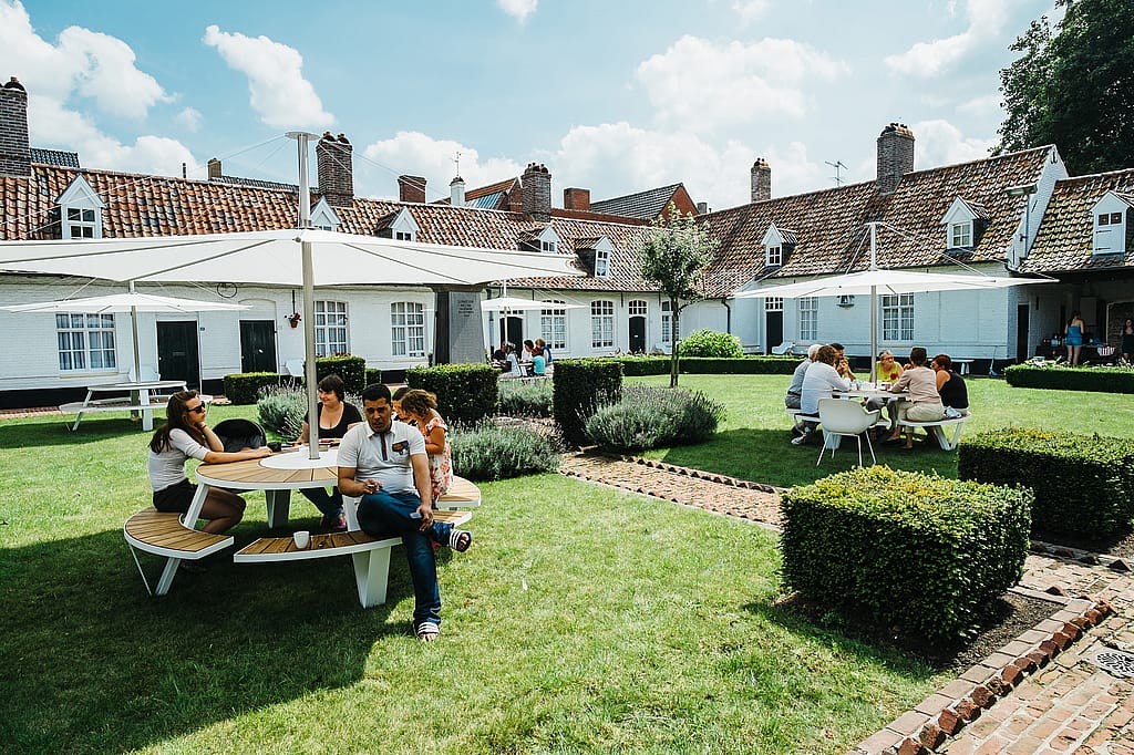 Several groups seated at Pantagruel tables with white parasols in a large grassy courtyard, with white brick buildings and tiled roofs in the background.