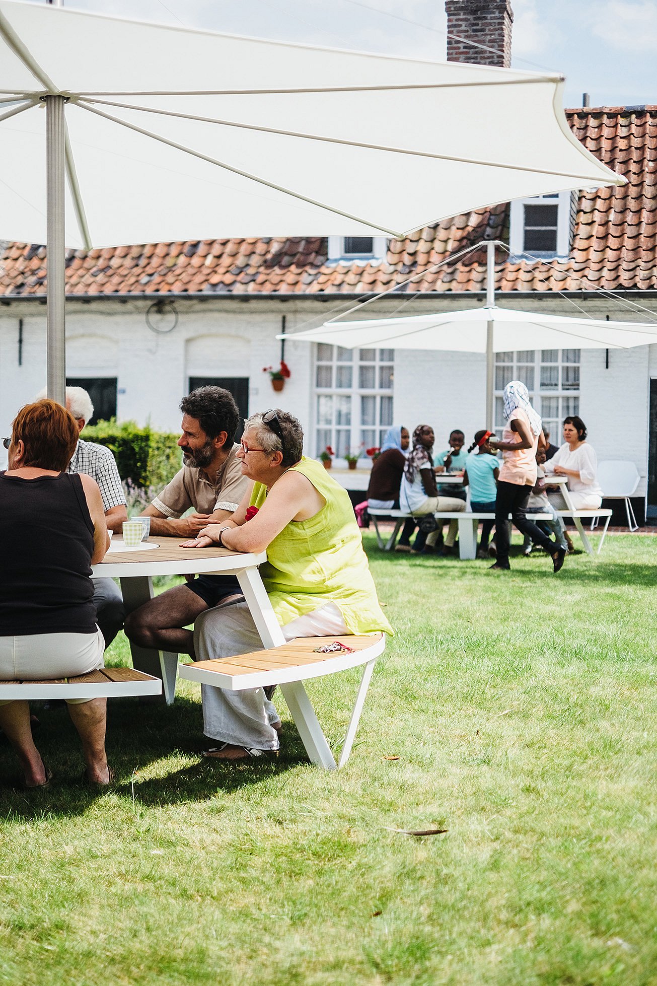 Close view of adults seated at a Pantagruel table under a white parasol, with more people gathered at another table in the background across the lawn.