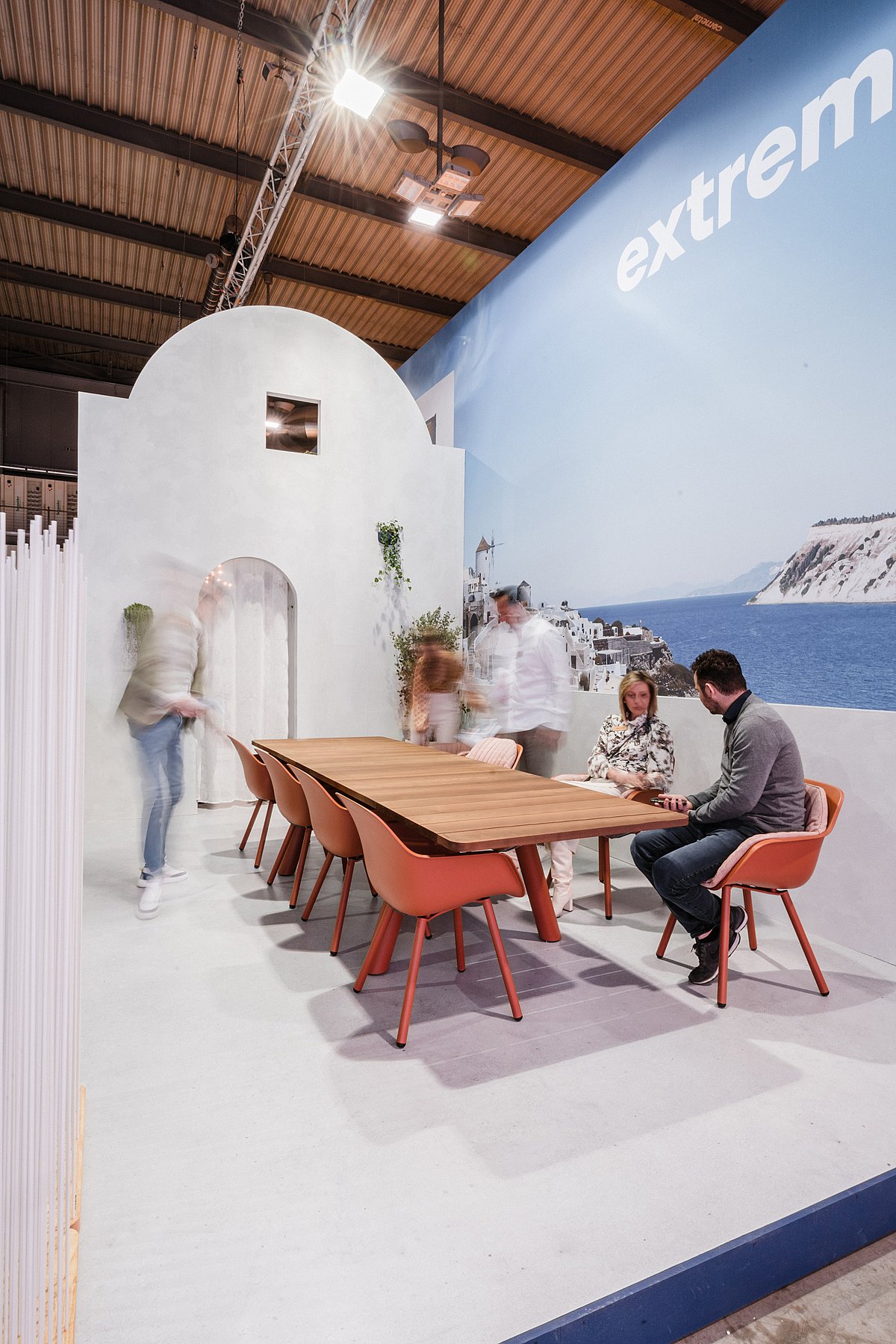 Lange houten Panigiri-tafel met terracotta stoelen in een witgekalkte, op Santorini geïnspireerde kermis, met een gewelfde deuropening en een muurschildering aan zee op de achtergrond.