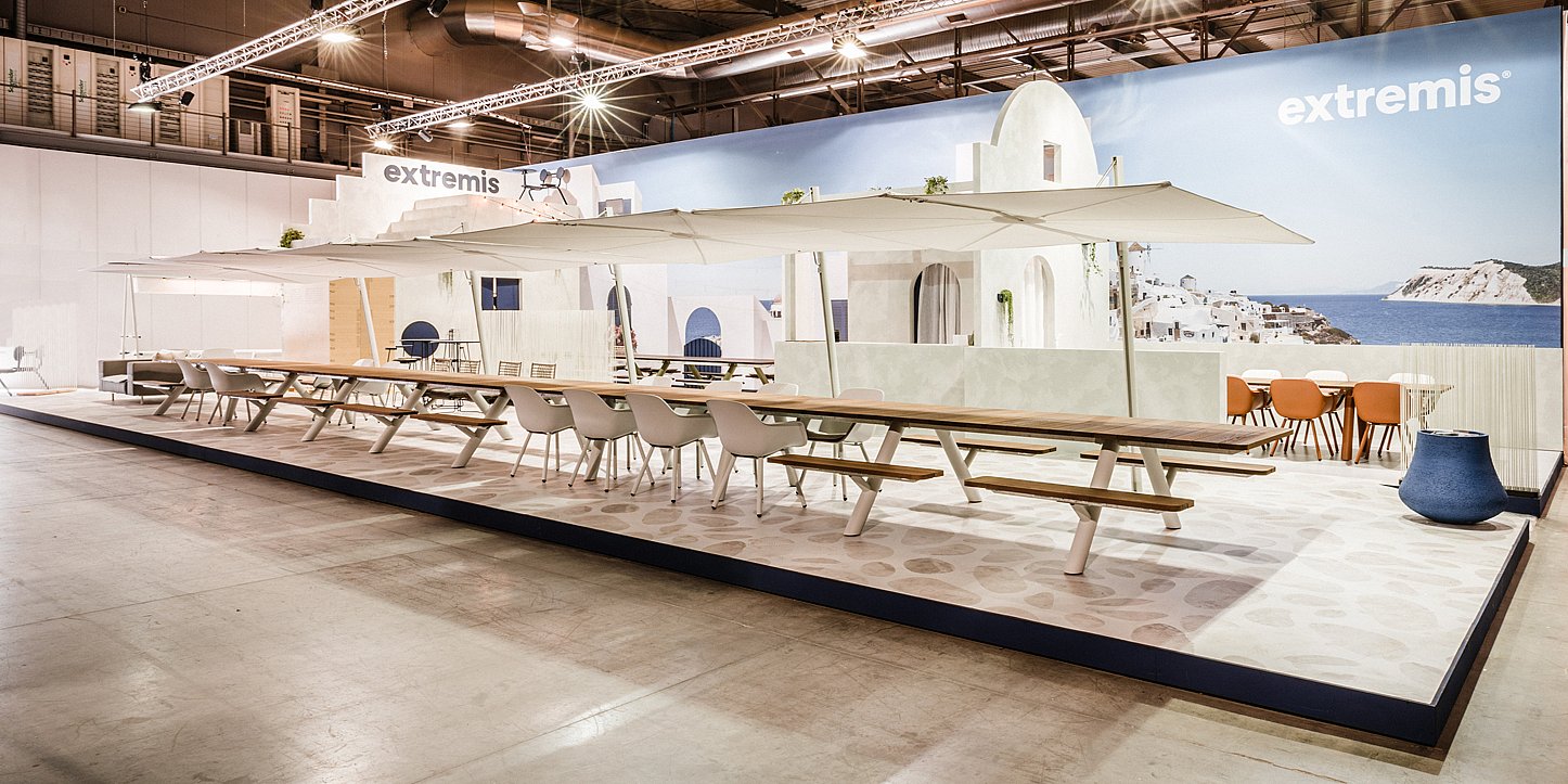 Breed zicht op de Extremis Salone-standaard met extra lange Panigiri-tafels, witte tinten en een blauwe muurschildering aan zee die de achterwand omhult.