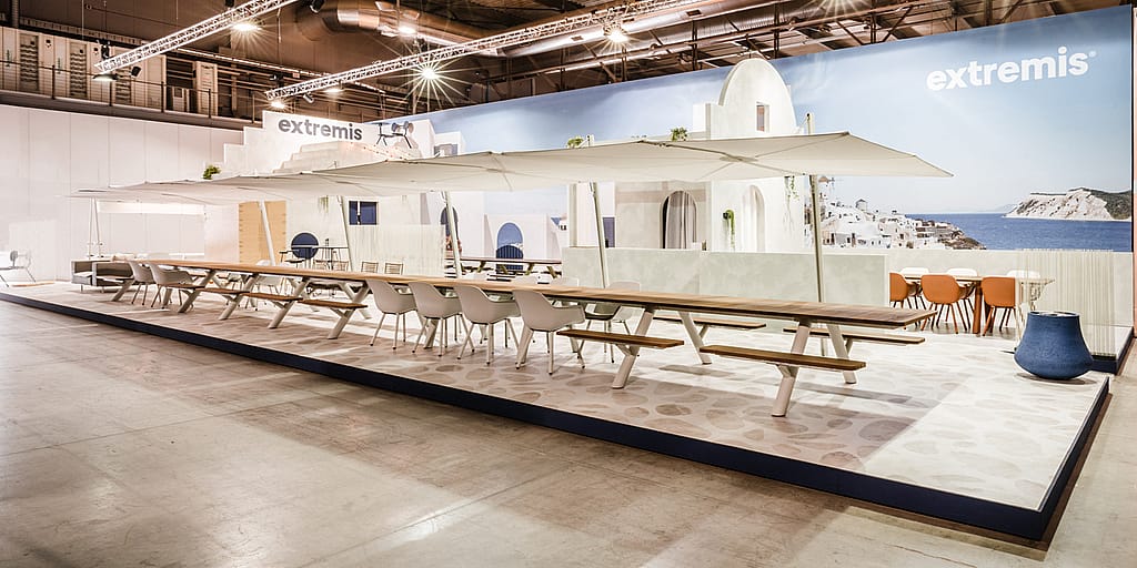 Breed zicht op de Extremis Salone-standaard met extra lange Panigiri-tafels, witte tinten en een blauwe muurschildering aan zee die de achterwand omhult.