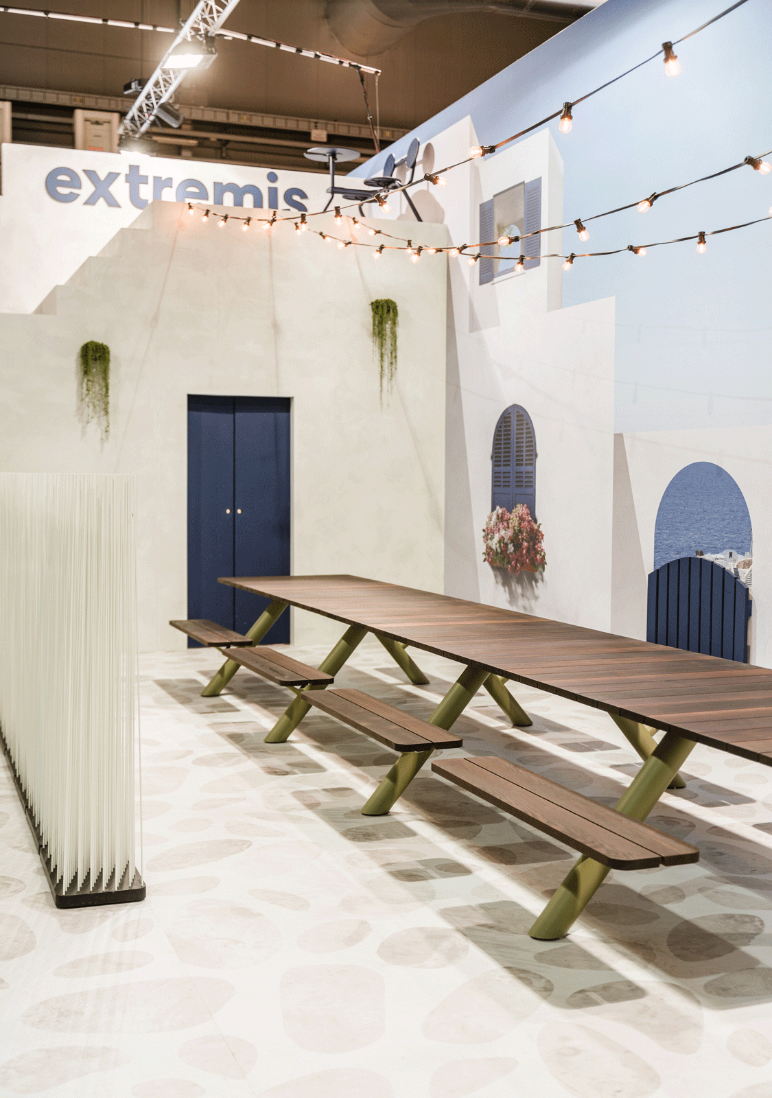 Lange houten Panigiri-tafel met terracotta kapiteinsstoelen in een witgekalkte, door Santorini geïnspireerde kermis, met een gebogen deuropening en een muurschildering aan zee op de achtergrond.