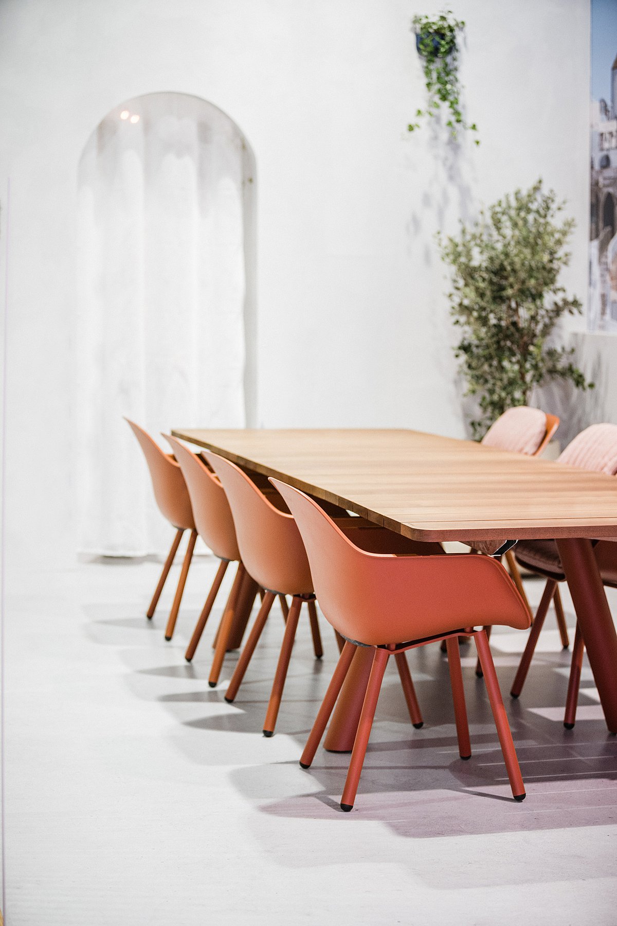 Dicht zijaanzicht van de Panigiri-tafel met het dikke houten blad en een rij terracotta kapiteinsstoelen in de witte tentoonstellingsomgeving.