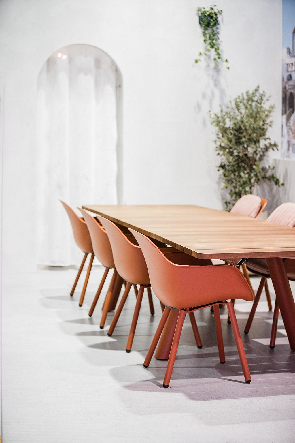 Dicht zijaanzicht van de Panigiri-tafel met het dikke houten blad en een rij terracotta kapiteinsstoelen in de witte tentoonstellingsomgeving.