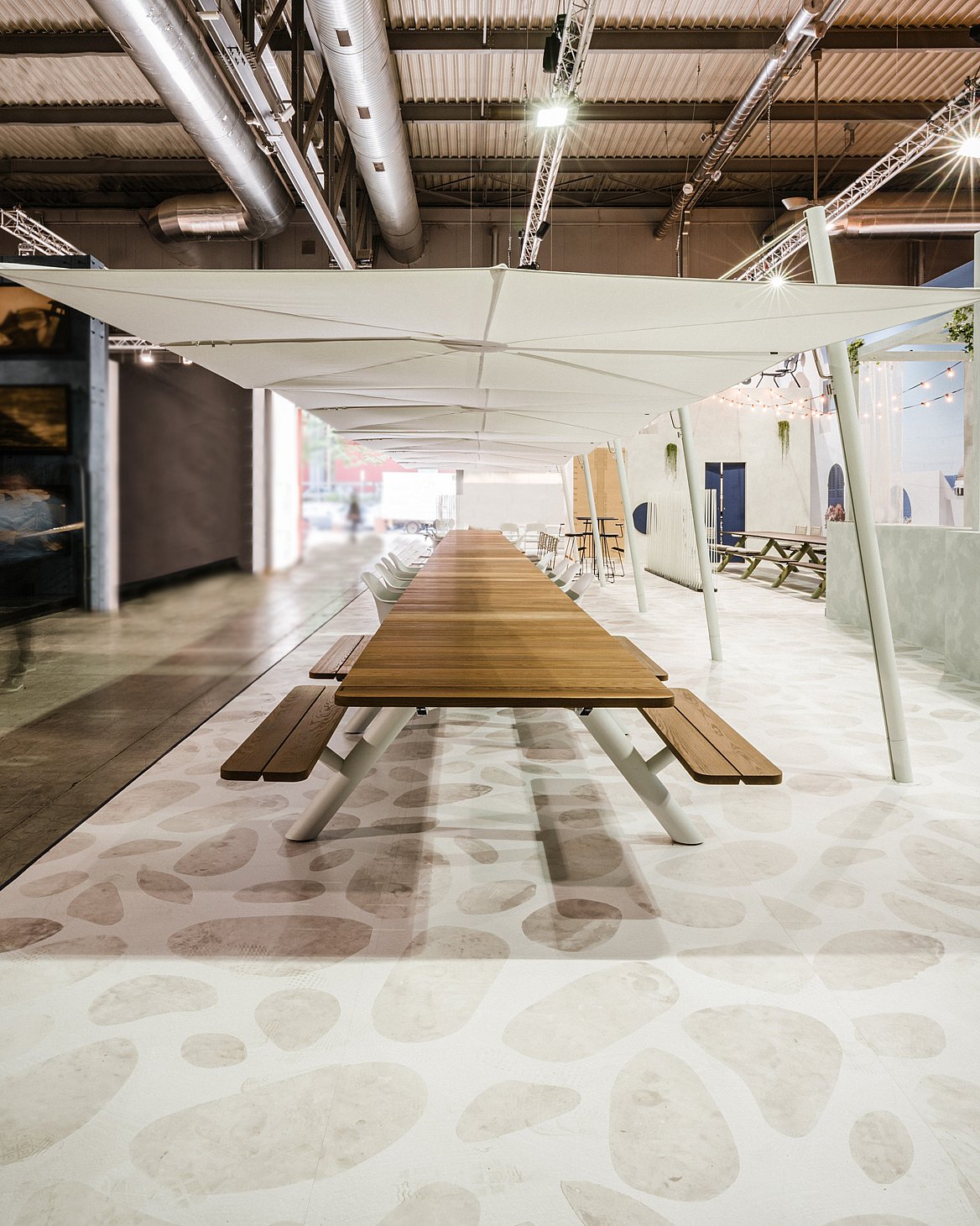 Gecentreerd uitzicht over de volledige lengte van een extra lange houten Panigiri-tafel met banken, die onder een doorlopende rij witte tinten loopt.