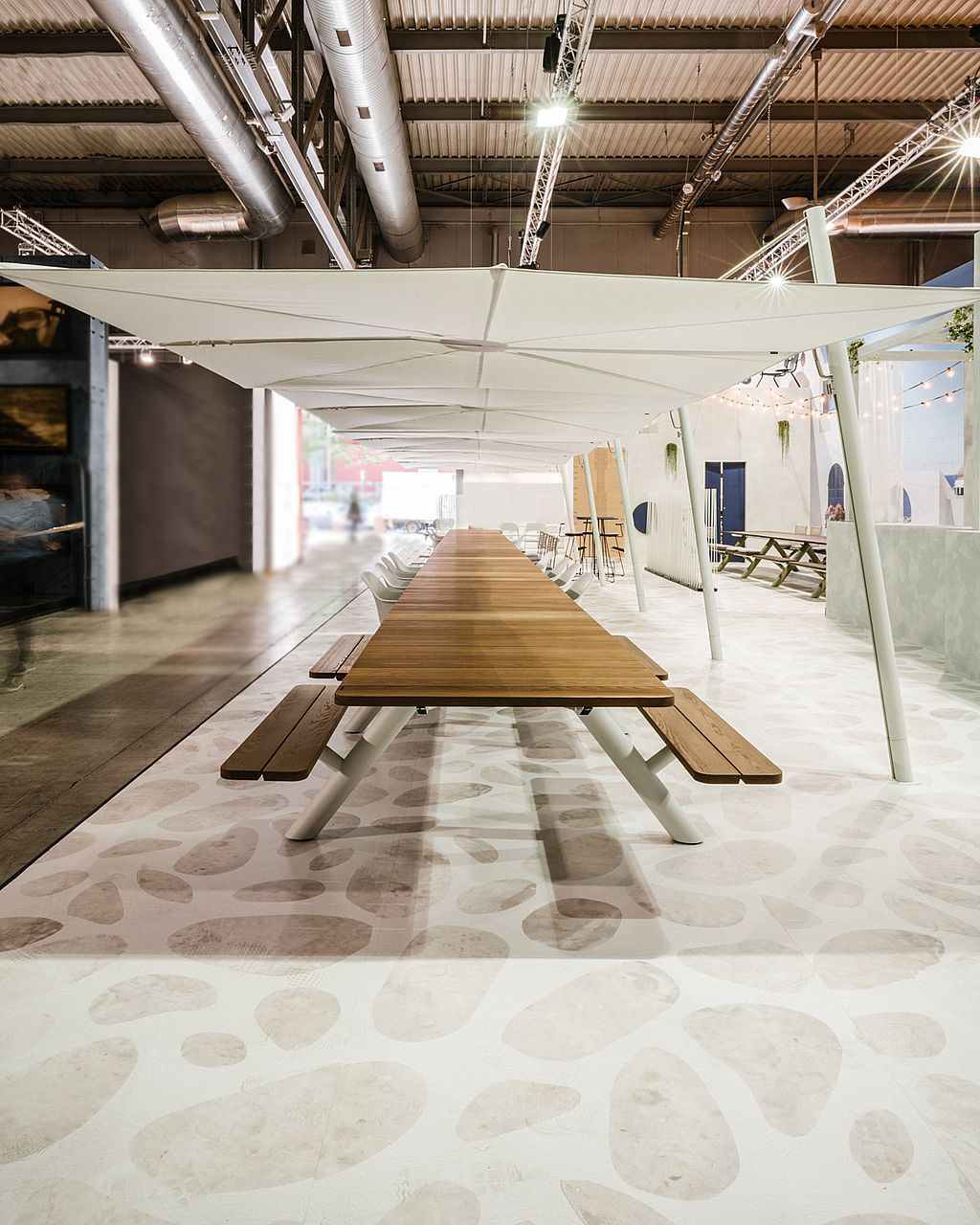 Gecentreerd uitzicht over de volledige lengte van een extra lange houten Panigiri-tafel met banken, die onder een doorlopende rij witte tinten loopt.