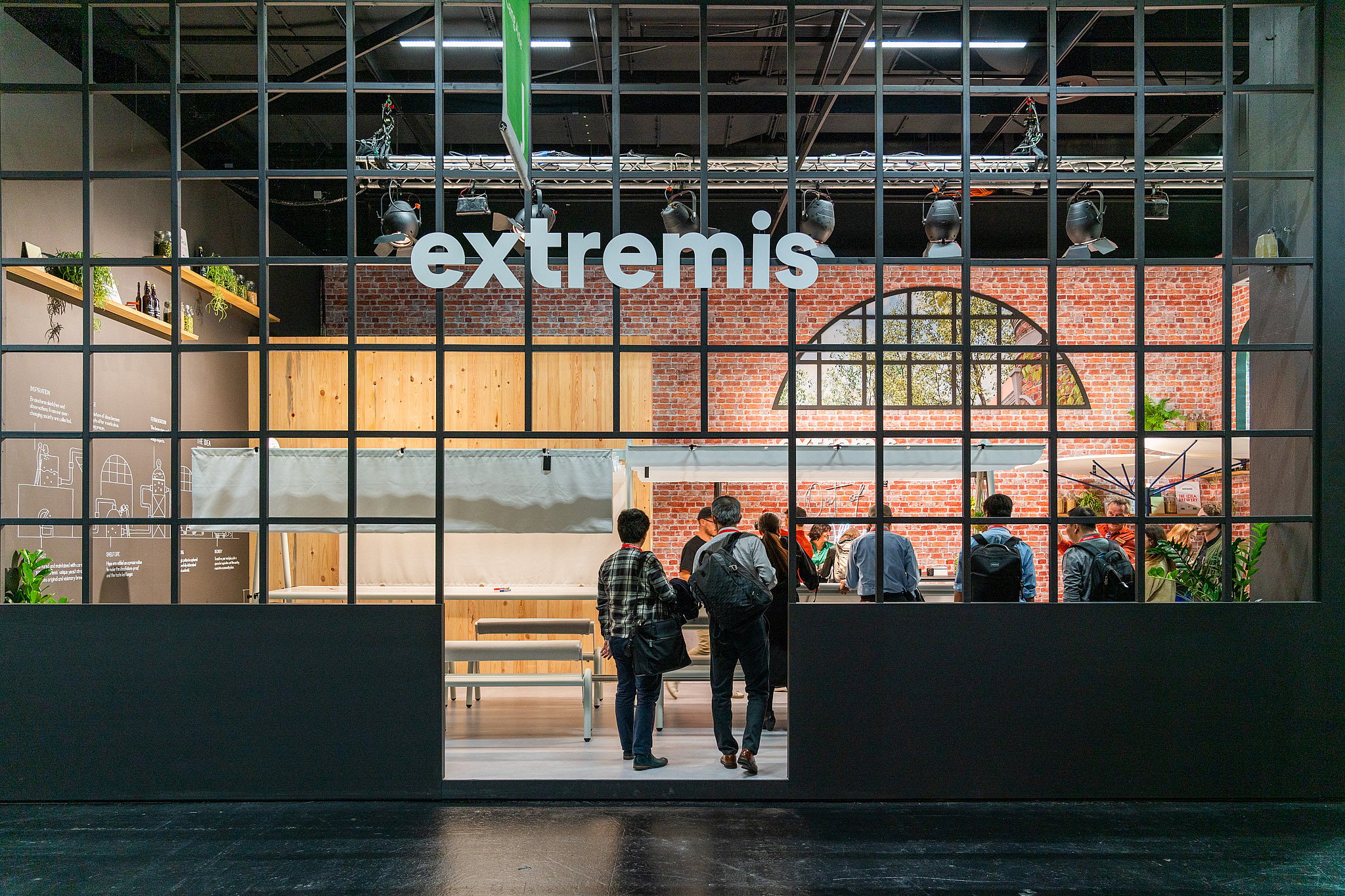 Vue de face du stand Extremis encadré par une façade en grille noire, avec les visiteurs entrant dans le stand inspiré de la brasserie et les murs en briques visibles à l’intérieur.