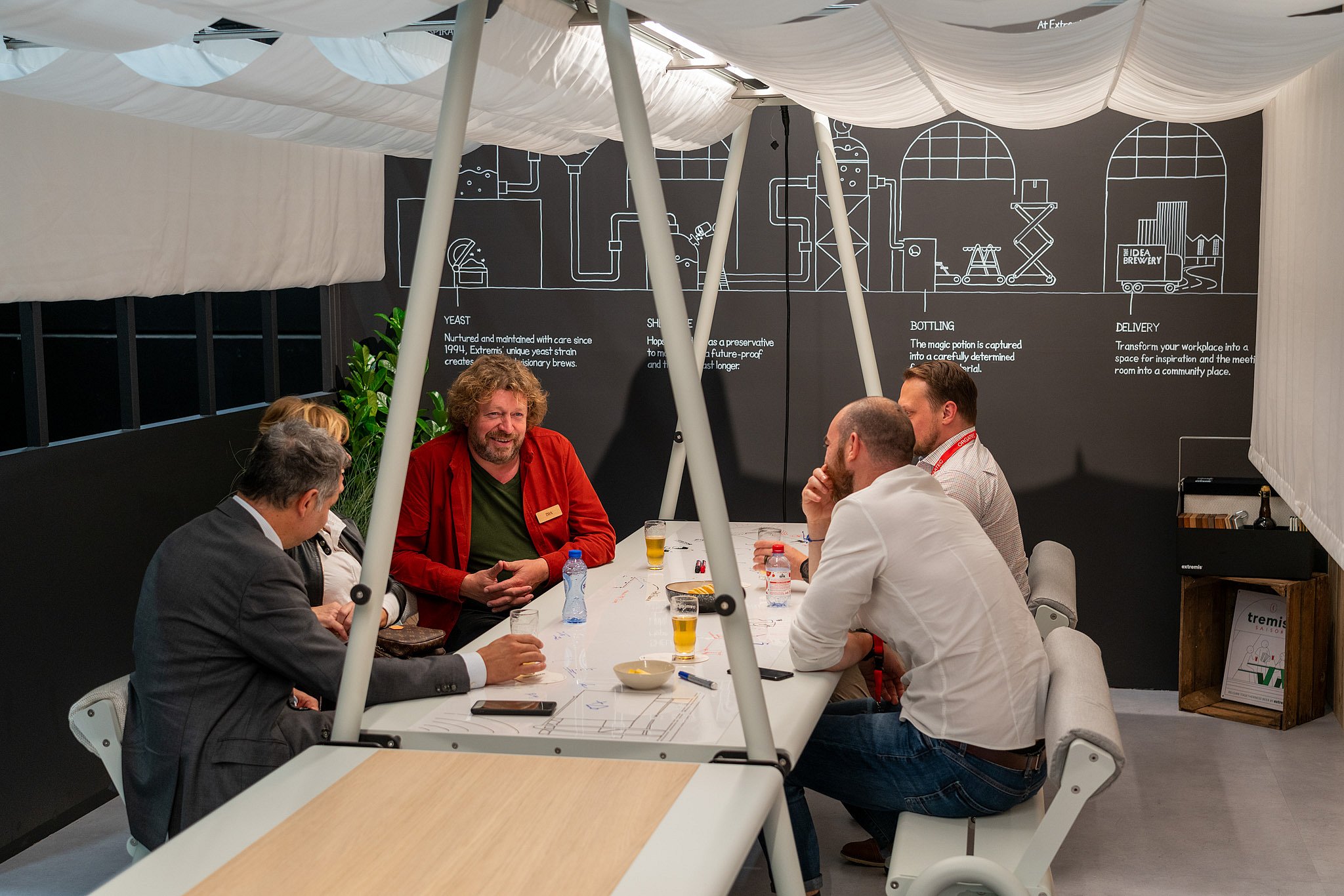 Cinq personnes assises autour d’une longue table blanche AMAi à l’intérieur d’un auvent acoustique, discutant autour d’un verre dans un décor d’exposition sur le thème de la brasserie.