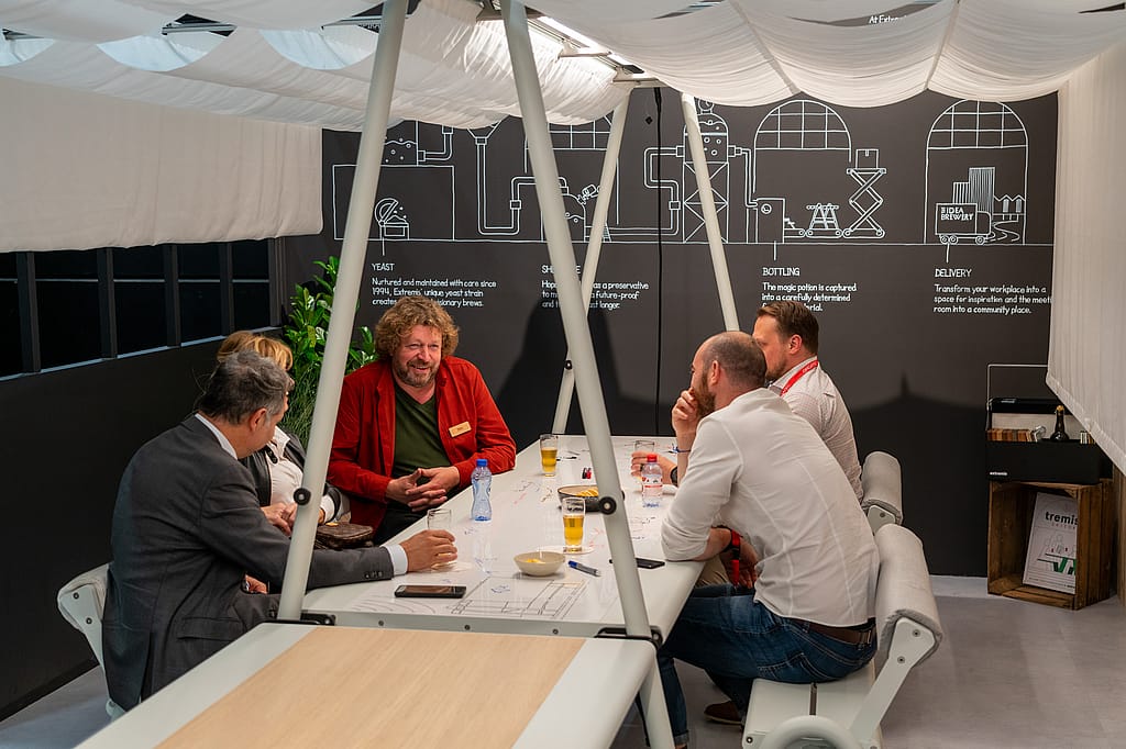 Fünf Personen sitzen um einen langen weißen AMAi-Tisch in einem akustischen Vordach mit Vorhängen und unterhalten sich bei Getränken in einer Ausstellungskulisse zum Thema Brauerei.