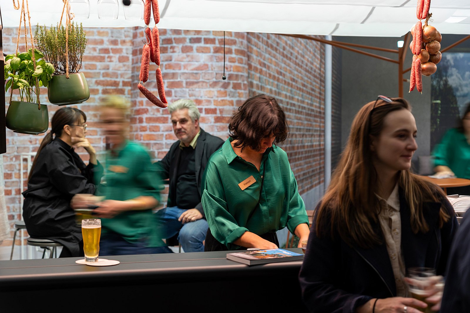 Scène de bar animée avec des saucisses suspendues et des herbes au-dessus, tandis que les visiteurs discutent et qu’un membre du personnel en vert organise le matériel derrière le comptoir de l’AMAi.