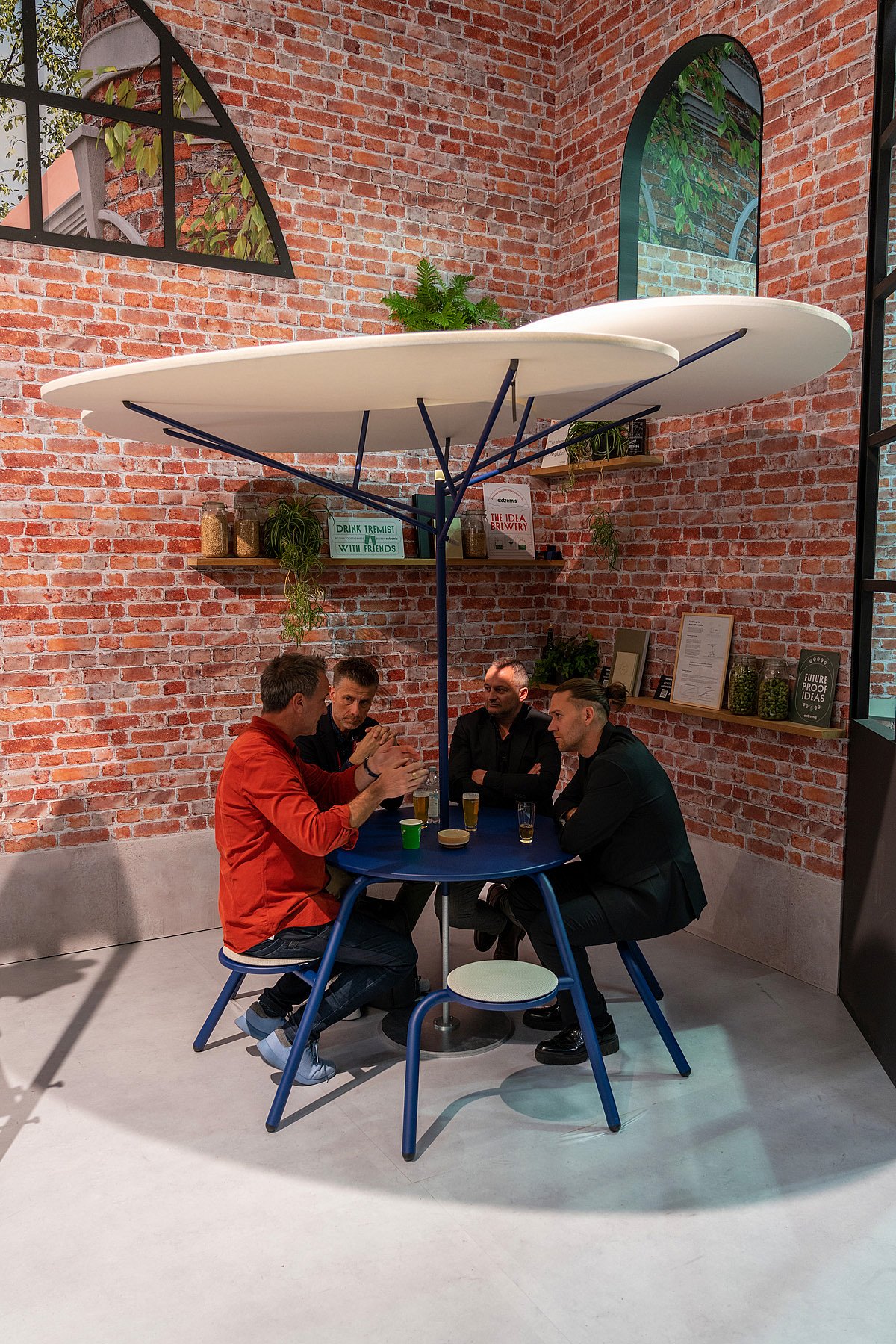 Quatre hommes assis autour d’une table haute ronde bleue sous une grande sculpture en acacia blanc dans un coin d’exposition aux murs de briques.