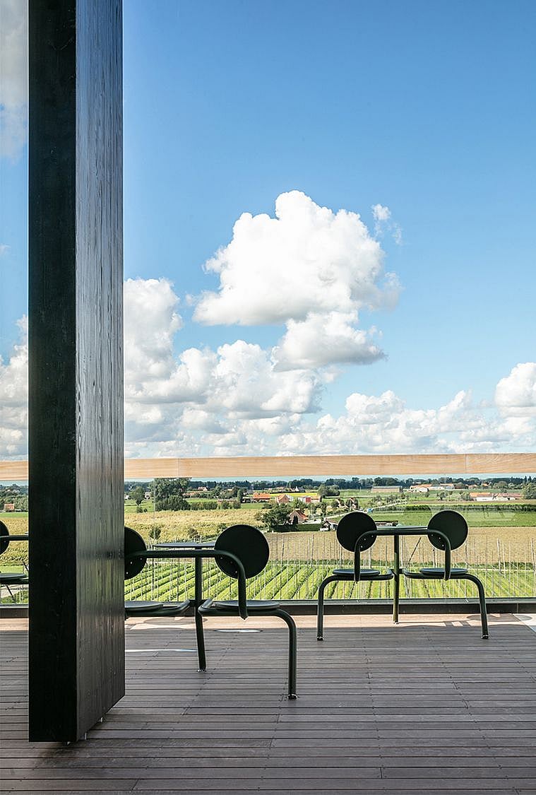 Paar grüne Bistrohocker mit kleinen runden Tischplatten, die entlang einer Glasbalustrade platziert sind und einen Blick auf die darunter liegenden Hopfenfelder bieten.