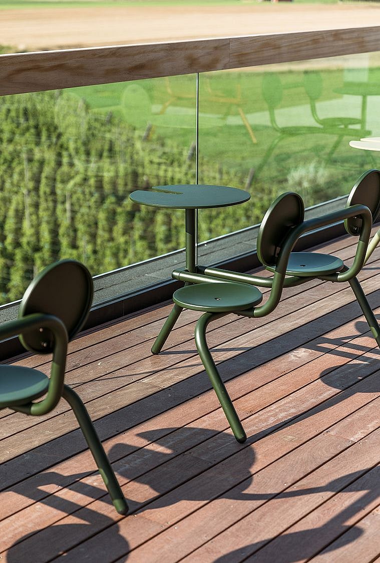 Paar grüne Bistrohocker mit kleinen runden Tischplatten, die entlang einer Glasbalustrade platziert sind und einen Blick auf die darunter liegenden Hopfenfelder bieten.