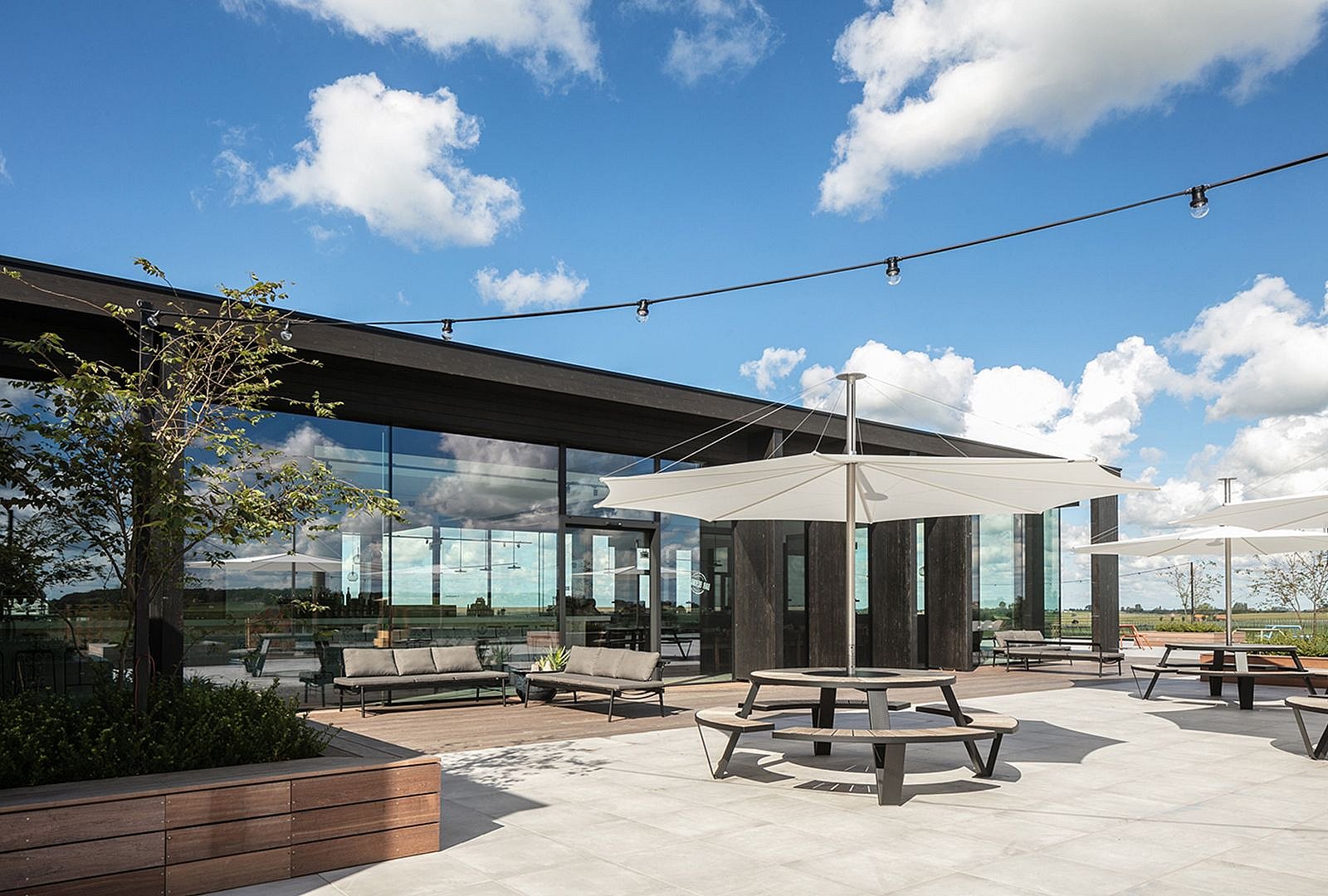 Helle Dachterrasse in der Bar Bernard mit verstreuten Tischen und Stühlen im Freien mit Blick auf offene Felder unter einem blauen Himmel mit Wolken.