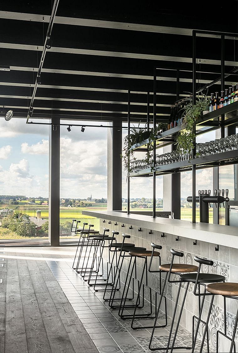 Paar schwarze Tiki-Hocker mit kleinen runden Tischplatten, die entlang einer Glasbalustrade platziert sind und einen Blick auf die darunter liegenden Hopfenfelder bieten.