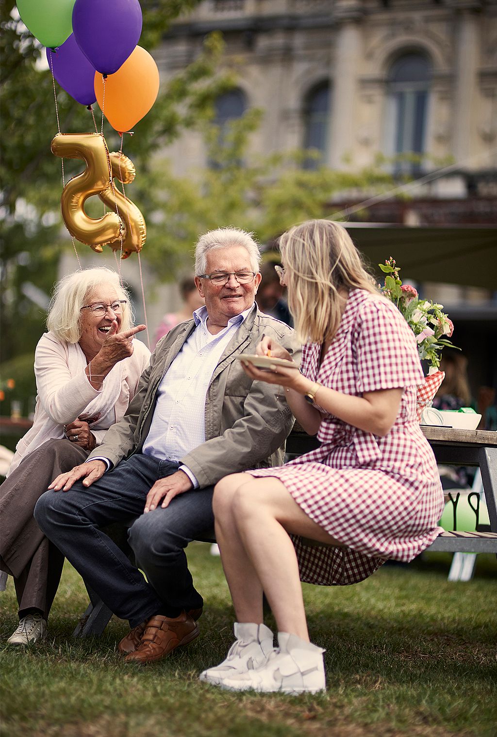 Ein älteres Paar lacht auf einer Bank neben bunten Luftballons in Form der Zahl „25“, während sich eine Frau im karierten Kleid mit ihnen unterhält.