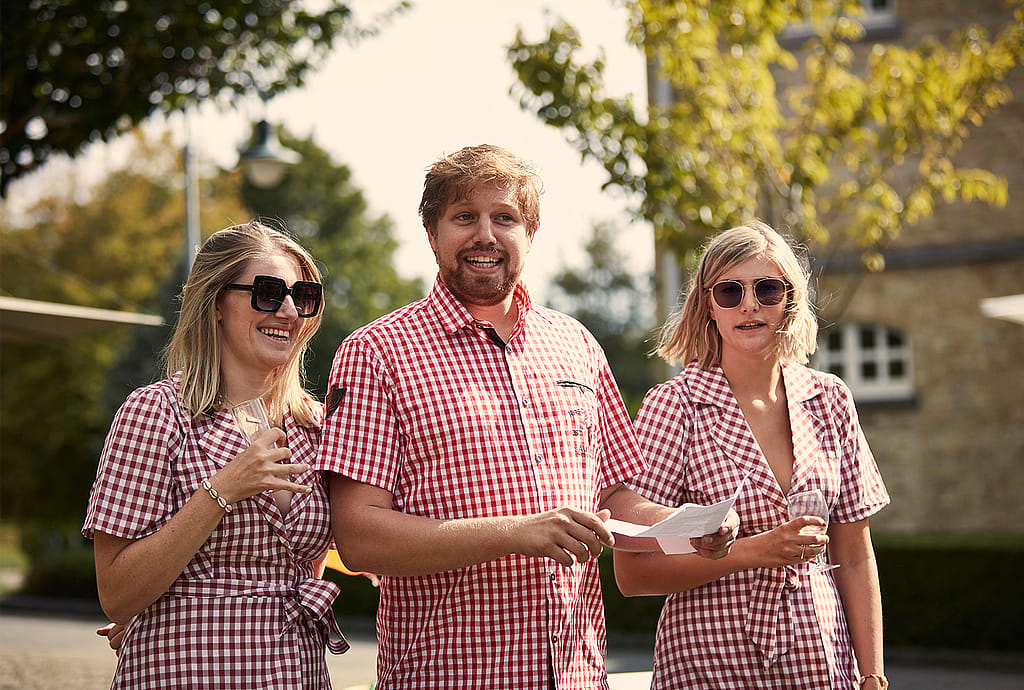 Die Geschwister Wynant in passenden rot-weiß karierten Outfits stehen während der Feier beisammen und halten Gläser und Papiere.