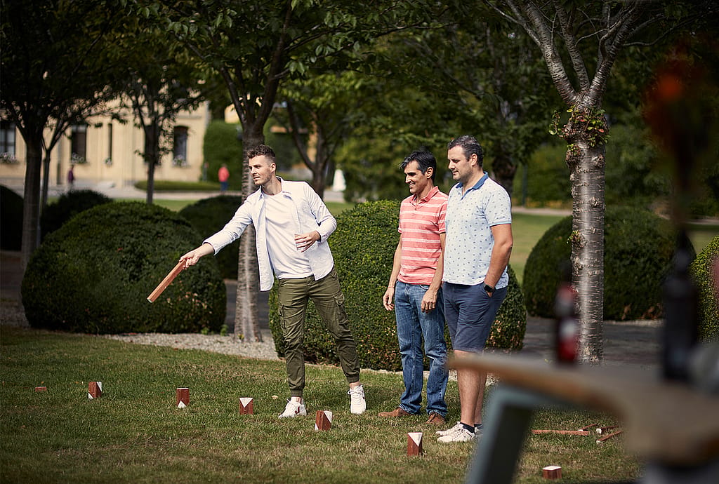 Drei Männer spielen ein Rasenspiel, bei dem sie einen Holzstab auf kleine Holzklötze im Gras werfen.