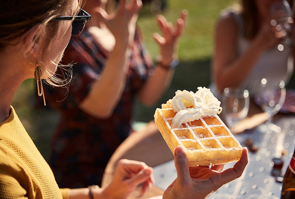 Eine Frau hält an einem Picknicktisch eine rechteckige, mit Sahne bedeckte Waffel hoch, während Freunde im Hintergrund gestikulieren.