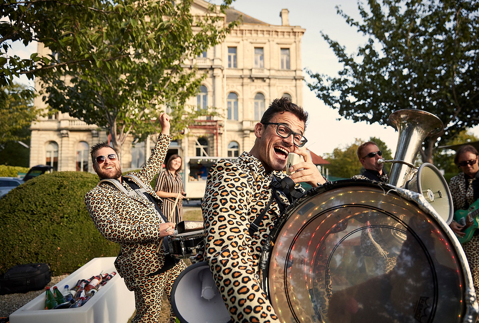 Ein lächelnder Musiker im Leopardenanzug deutet auf die Kamera, während er eine beleuchtete Basstrommel spielt; seine Bandkollegen stehen hinter ihm.
