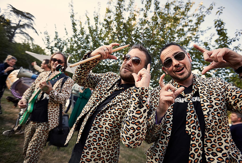 Bandmitglieder in Leopardenanzügen posieren mit Instrumenten und verspielten Handgesten während der Gartenparty.