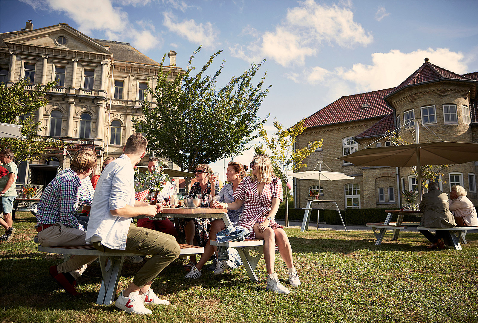 An einem sonnigen Nachmittag sitzen Freunde um einen runden Gargantua-Tisch auf der Wiese zwischen historischen Gebäuden und großen Sonnenschirmen.