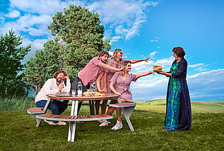 Die Familie Wynant sitzt an einem runden Gargantua-Tisch und streckt eifrig die Hände nach der Mutter aus, die einen Stapel Waffeln trägt. Der strahlend blaue Himmel bildet den Hintergrund, während der Vater mit einem Getränk daneben sitzt.