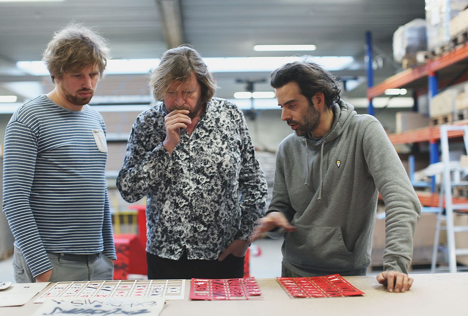 Denis Meyers se tient avec deux autres personnes autour d’une table de travail, discutant des maquettes rouges imprimées ou peintes dans l’atelier.