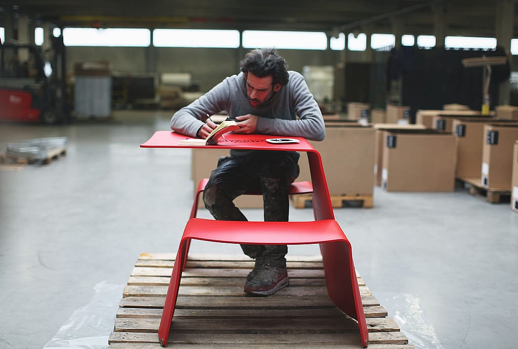 Denis Meyers est assis à une simple table Picnik rouge sur une palette en bois dans un atelier, peignant à la main des lettres noires sur le dessus de la table