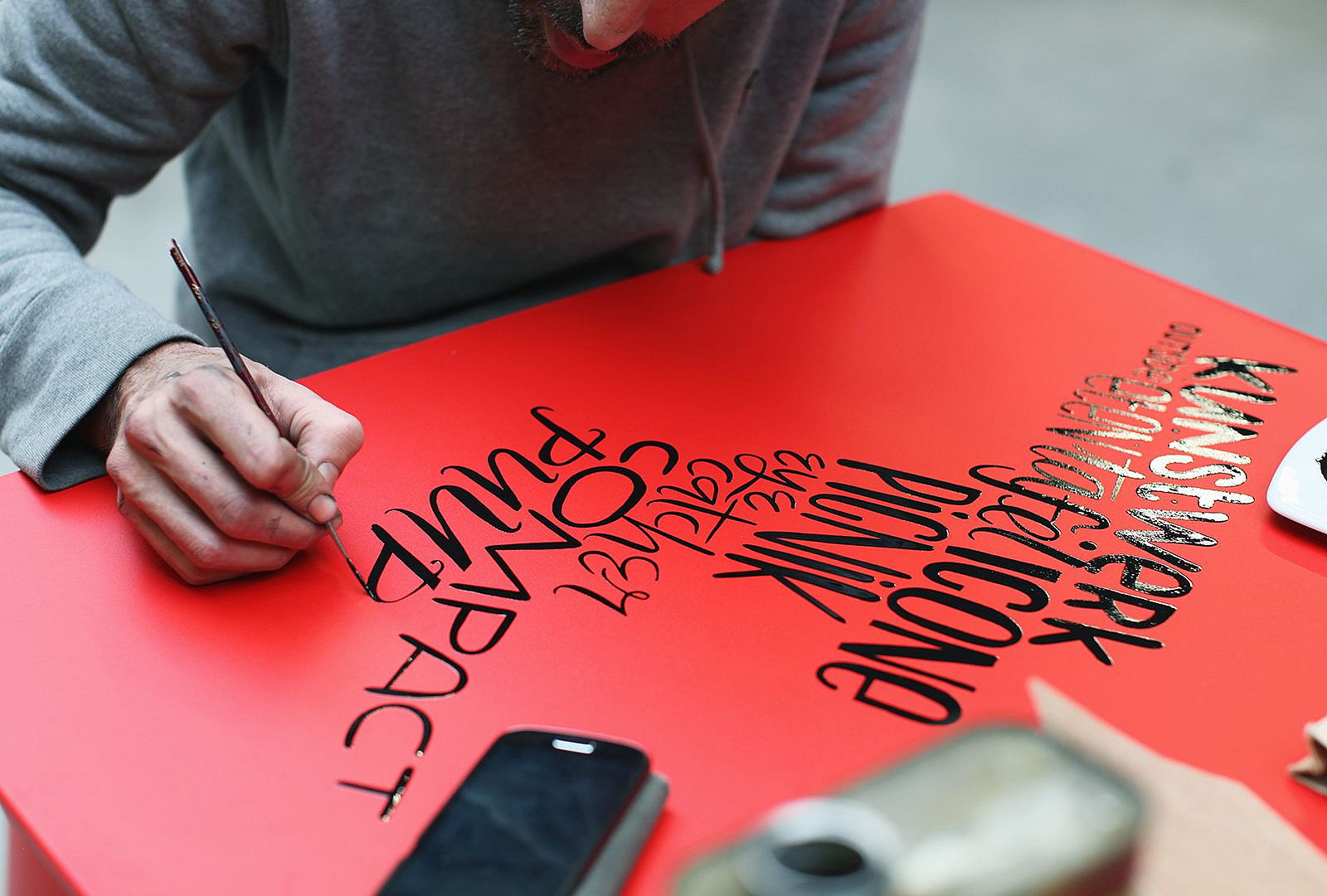 Denis Meyers est assis à une simple table de Picnik rouge sur une palette en bois dans un atelier, peignant à la main des lettres noires sur le plateau.