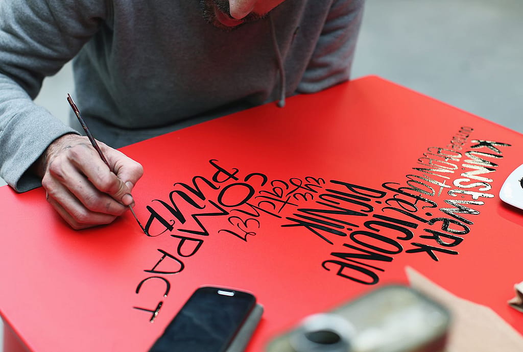Denis Meyers est assis à une simple table de Picnik rouge sur une palette en bois dans un atelier, peignant à la main des lettres noires sur le plateau.