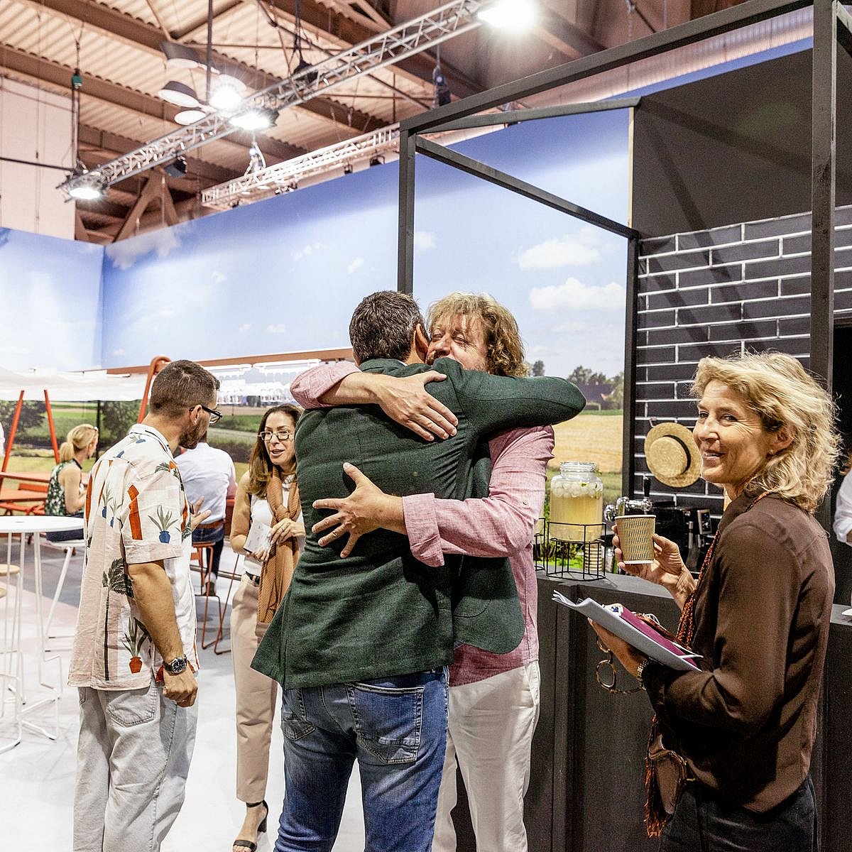 Les gens s’étreignent et discutent à côté de l’espace bar du stand, capturant le sentiment chaleureux des retrouvailles dans la vraie vie.