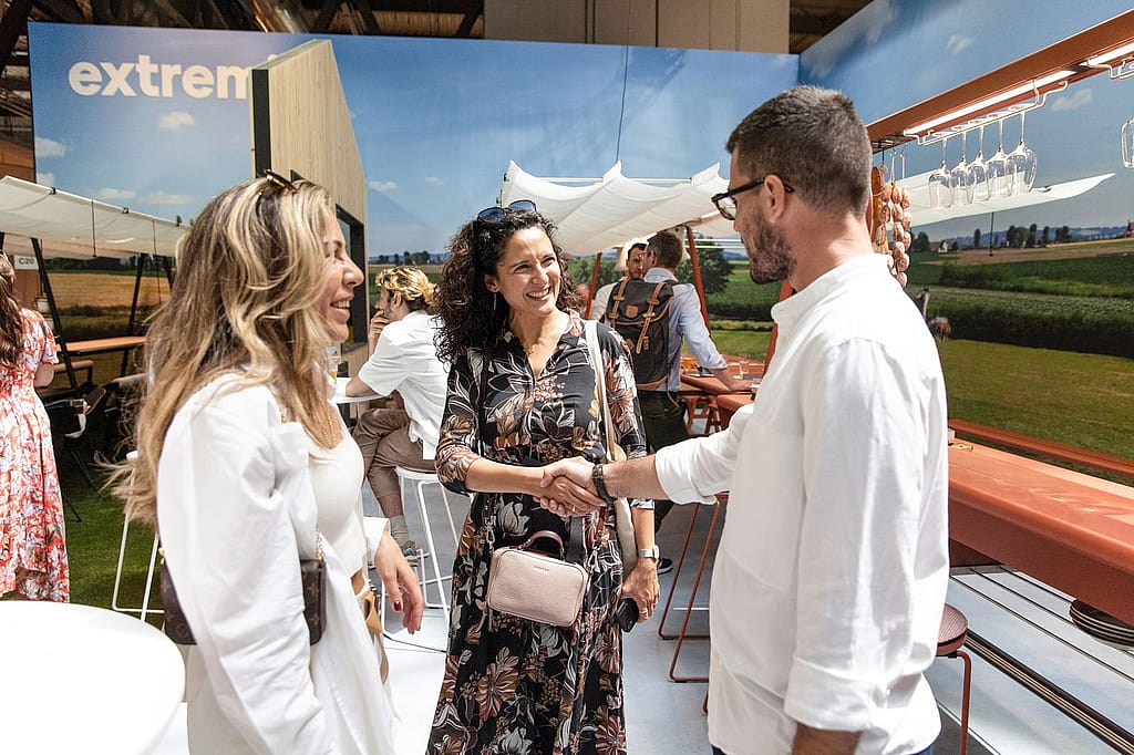 Trois visiteurs se saluent à l’intérieur du stand d’Extremis à Milan, serrant la main et souriant devant une fresque murale de campagne et des meubles en terre cuite.
