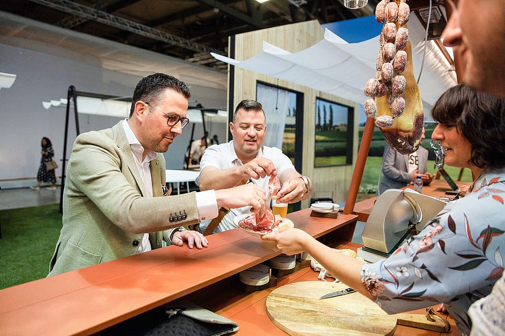 Deux hommes reçoivent des boissons et des collations à travers un comptoir de bar de couleur terre cuite, avec des charcuteries suspendues et une desserte en bois au premier plan.