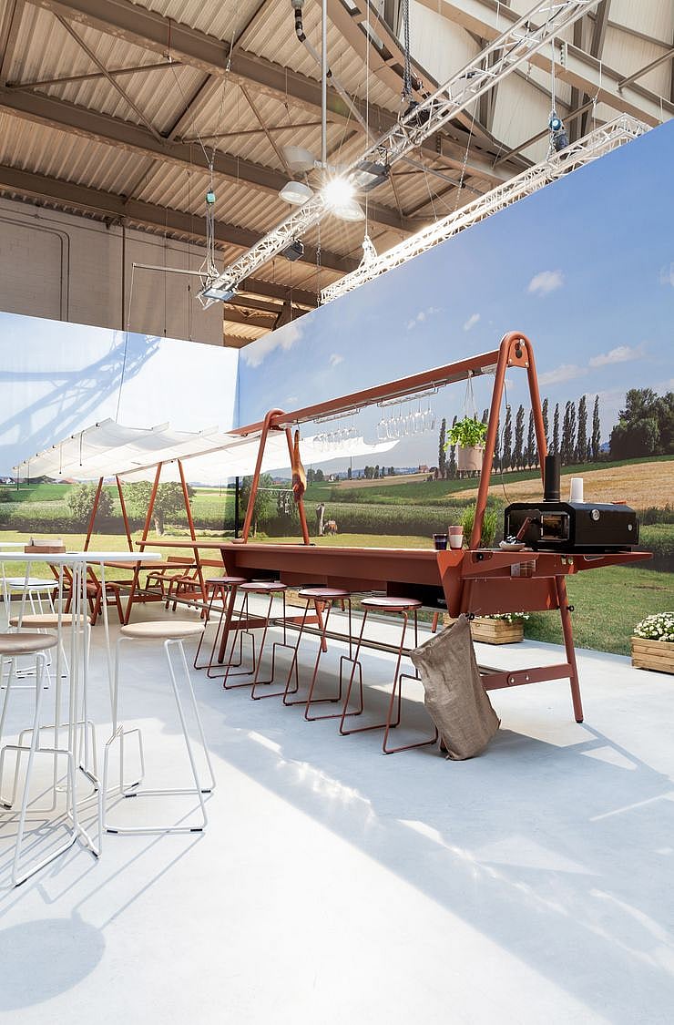 Vue complète du stand montrant l’installation modulaire AMAi disposée comme une plate-forme de jardin avec de longues tables, des tabourets et un auvent en tissu blanc au-dessus.